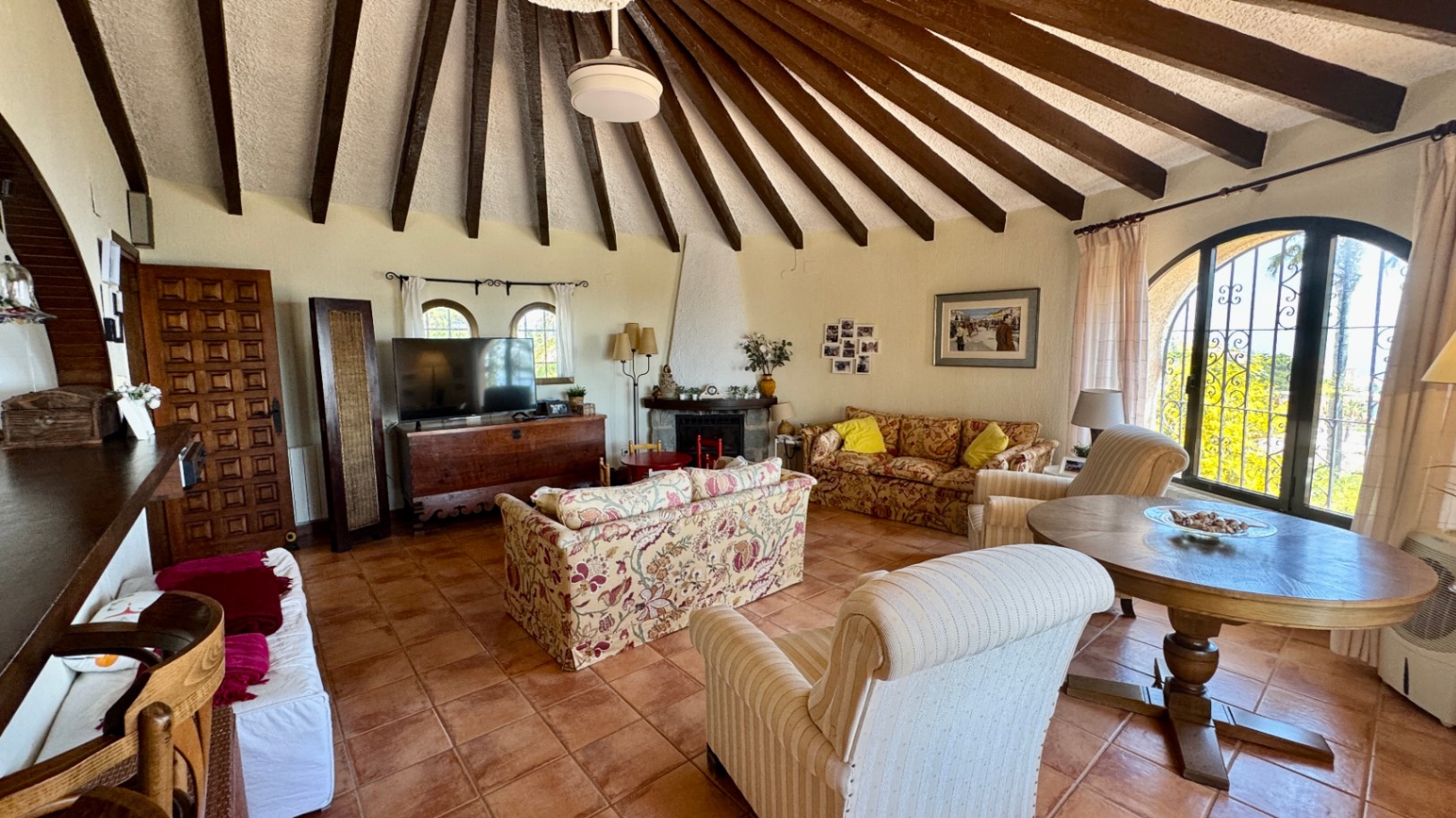 Ruime woonkamer met een gewelfd plafond met radiale houten balken, terracotta vloertegels, een open haard en boogramen met smeedijzeren decoratie.