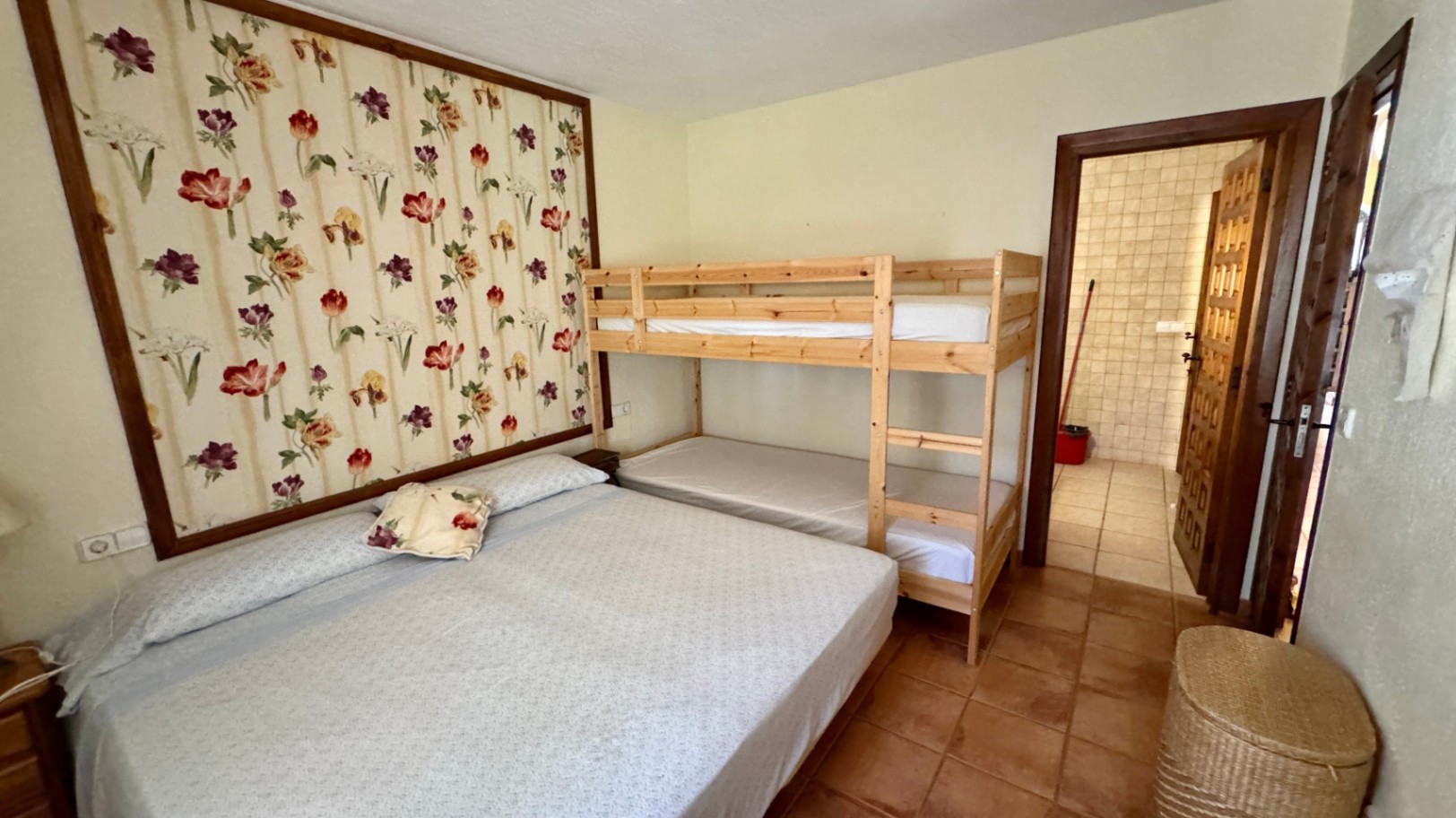 Slaapkamer met een tweepersoonsbed en houten stapelbed, voorzien van een bloemenbehangpaneel en terracotta vloertegels. Badkamer zichtbaar via de open deur.