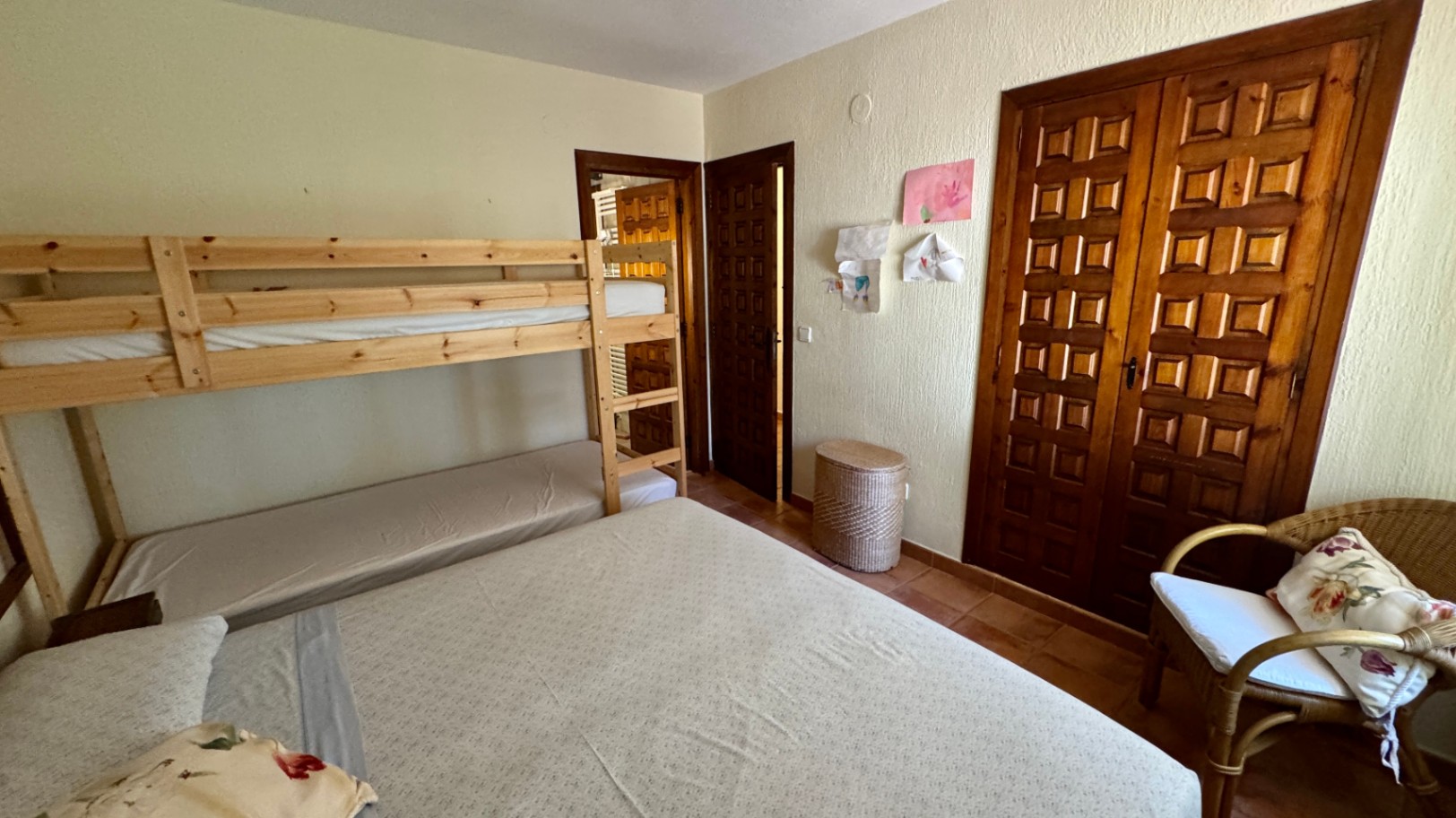 Slaapkamer met grenen stapelbedden, een ingebouwde houten kast met paneeldeuren en een terracotta tegelvloer. Voorzien van een rieten stoel en structuurwanden.