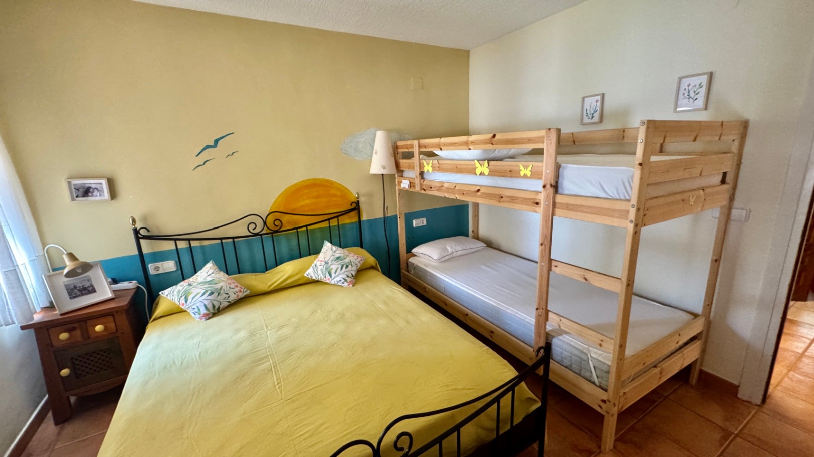 Slaapkamer voorzien van een tweepersoonsbed met smeedijzeren hoofdbord en een houten stapelbed. De ruimte heeft een terracotta tegelvloer en een muurschildering.