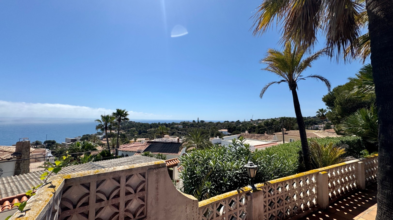 Verhoogd terrasuitzicht over een mediterrane woonwijk met terracotta daken, palmbomen en een verre zeehorizon onder een strakblauwe lucht.