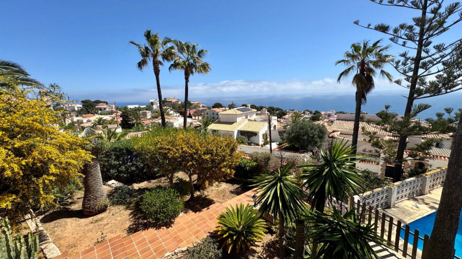 Verhoogd uitzicht op een mediterrane tuin met terracotta paden en palmbomen, uitkijkend over een residentiële kustwijk en de open zee.
