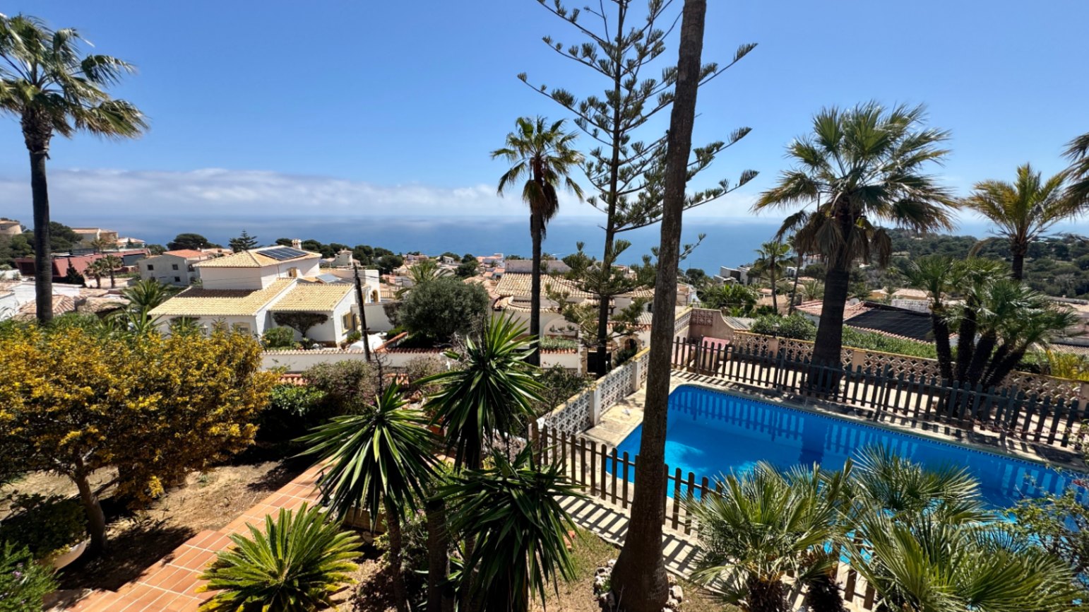 Verhoogd uitzicht op een privézwembad met houten hekwerk, omgeven door palmbomen en mediterrane tuinen met de zee aan de horizon.