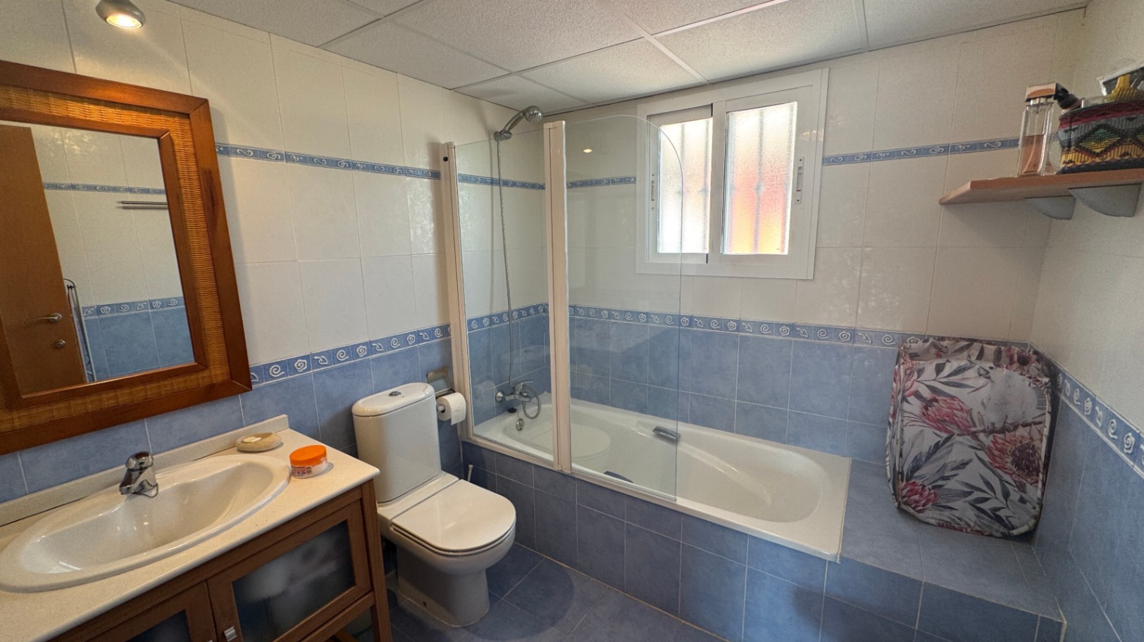 Badkamer met blauw-witte betegeling, een ligbad met glazen douchescherm, een spiegel met houten lijst en een bijpassend wastafelmeubel.