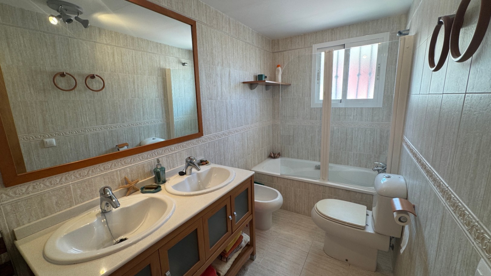Badkamer met dubbele wastafel op een houten meubel, een ligbad met glazen wand en wanden volledig betegeld in neutrale tinten met een sierrand.