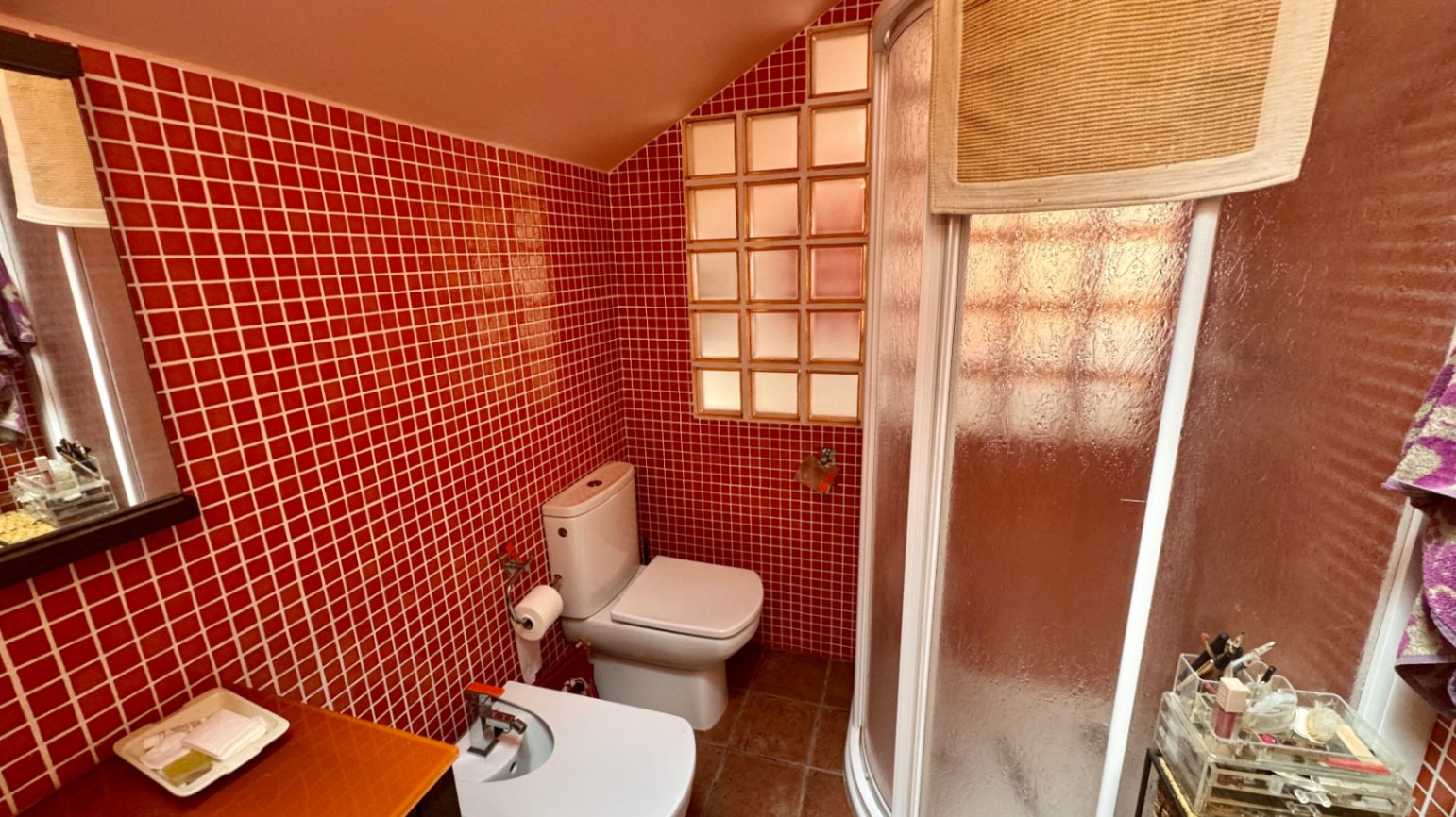 Badkamer met rode mozaïektegels, een schuin plafond en een halfronde hoekdouche. Voorzien van een toilet, bidet en een raam van glasbouwstenen.
