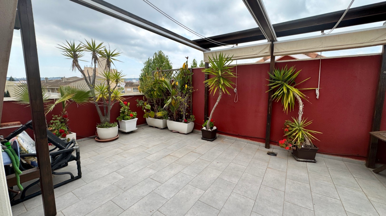 Ruim dakterras met grijze tegels, een houten pergola met uitschuifbare zonwering en diverse potplanten tegen een dieprode buitenmuur.