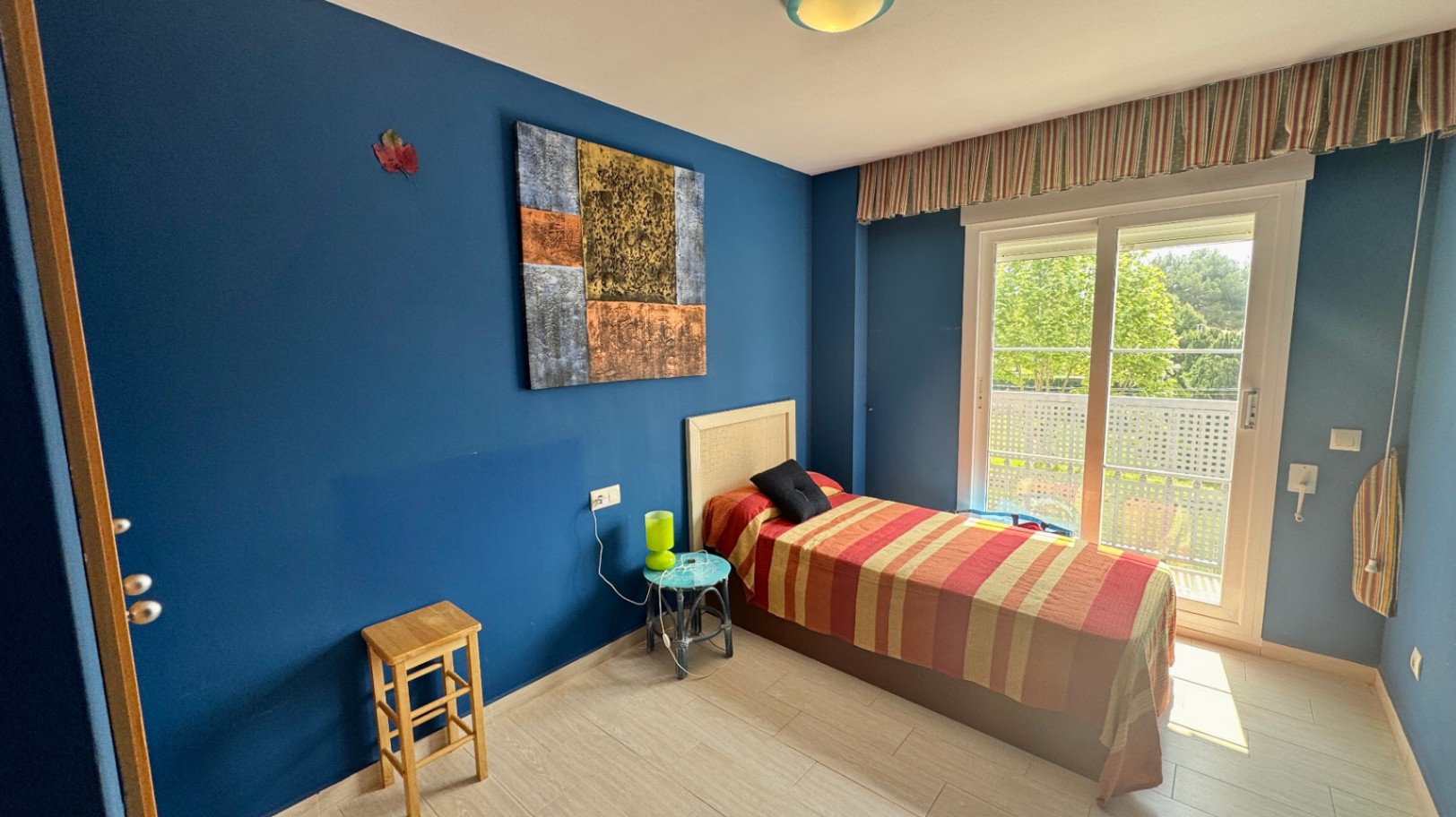 Slaapkamer met diepblauwe muren, een bed met rieten hoofdbord en een grote schuifpui die toegang biedt tot een privébalkon met uitzicht op de tuin.