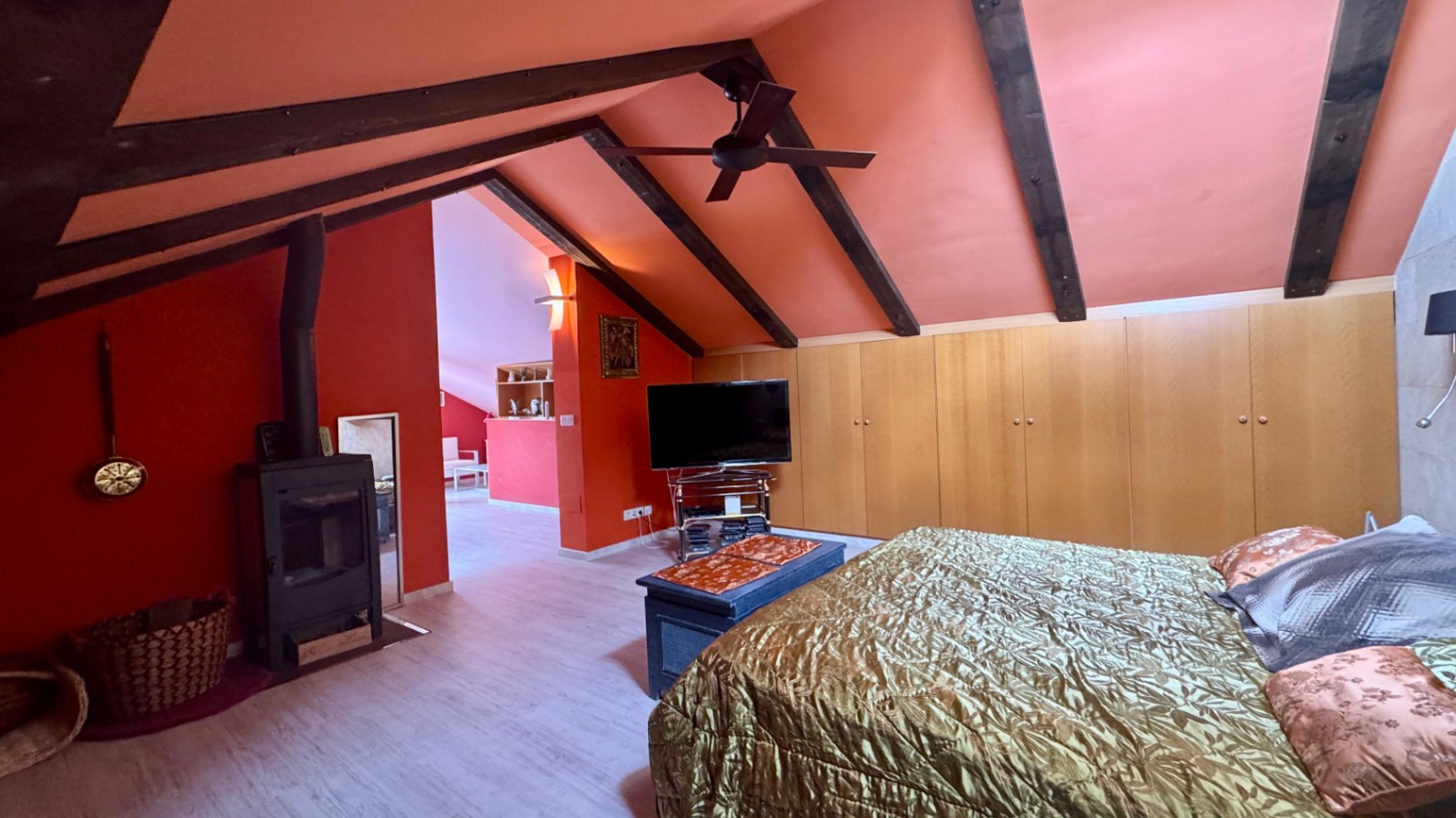 Slaapkamer onder het schuine dak met donkere houten balken, terracotta wanden, een houtkachel en kamerhoge houten inbouwkasten.
