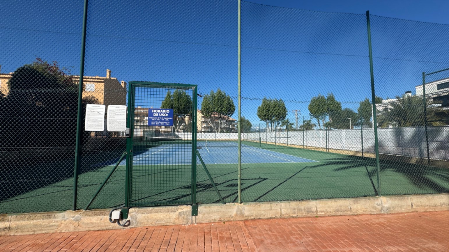 Buiten tennisbaan met een blauw en groen hardcourt oppervlak, volledig omheind door een hoog hekwerk met een afsluitbare metalen toegangspoort.