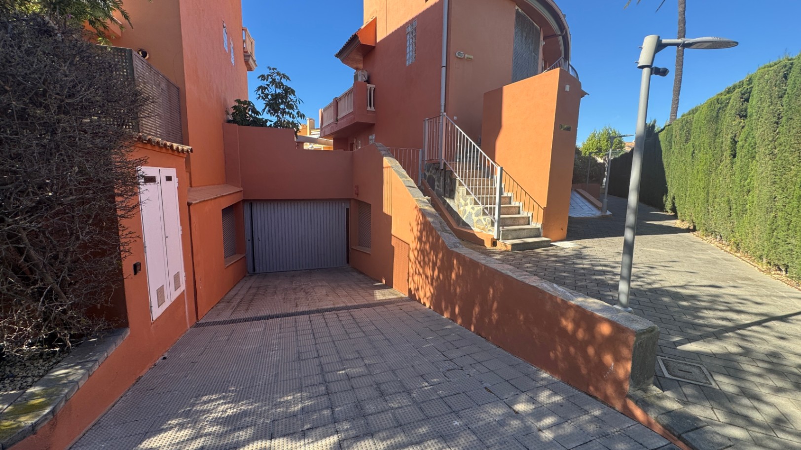Buitenaanzicht met een geplaveide oprit naar een privégarage, terracotta gevel en een buitentrap met metalen leuning naast een hoge haag.