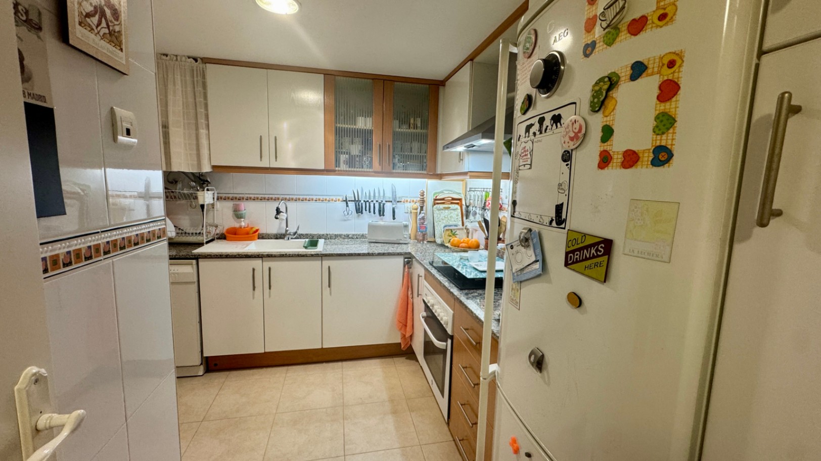 Functionele L-vormige keuken met witte kasten, houten accenten, een granieten werkblad en diverse inbouwapparatuur onder heldere verlichting.