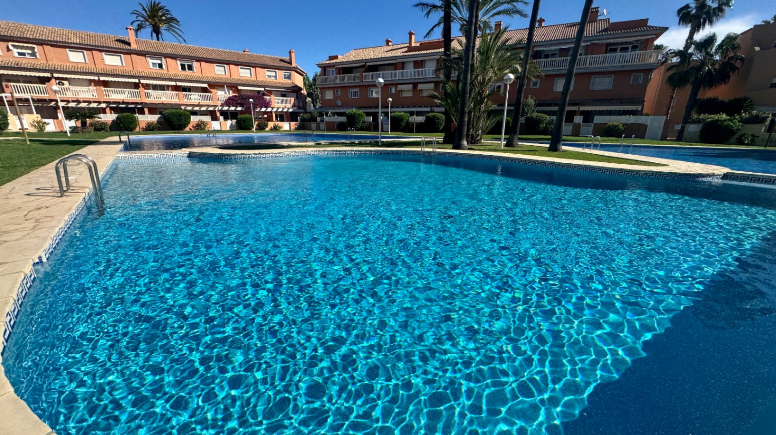 Groot gemeenschappelijk zwembad met blauwe mozaïektegels en een stenen rand, omgeven door aangelegde tuinen en mediterrane woongebouwen.