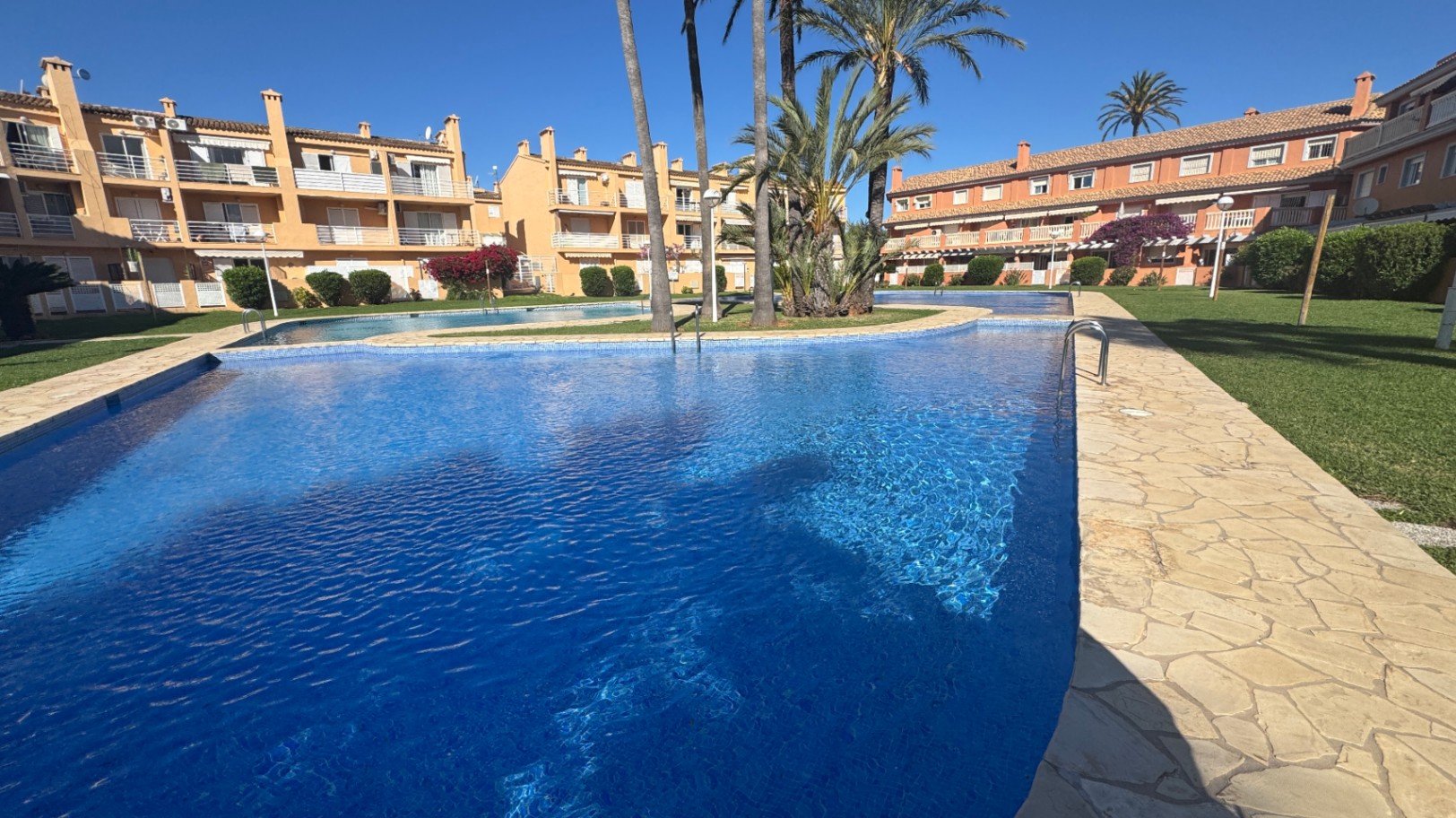 Groot gemeenschappelijk zwembad met blauwe mozaïektegels en natuurstenen randen, omgeven door gazons, palmbomen en mediterrane appartementen.