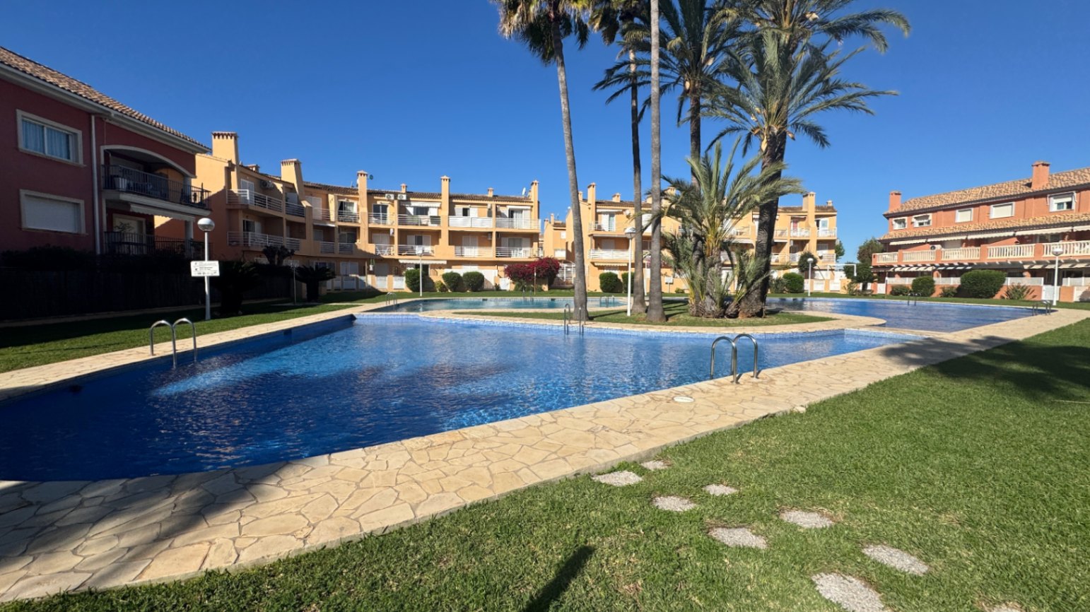 Groot gemeenschappelijk zwembad met blauwe tegels, omgeven door een stenen zonneterras, verzorgde gazons en palmbomen in een mediterrane residentie.