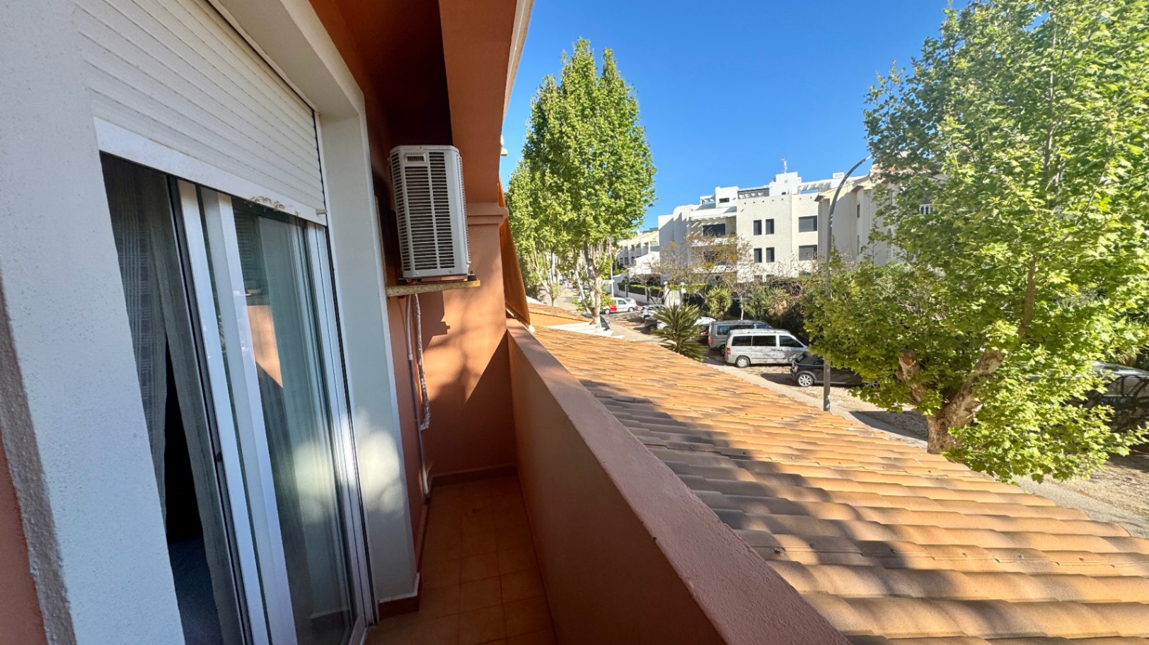 Smal balkon met terracotta tegels, een buitenunit voor airconditioning en uitzicht op een boomrijke straat met woongebouwen.