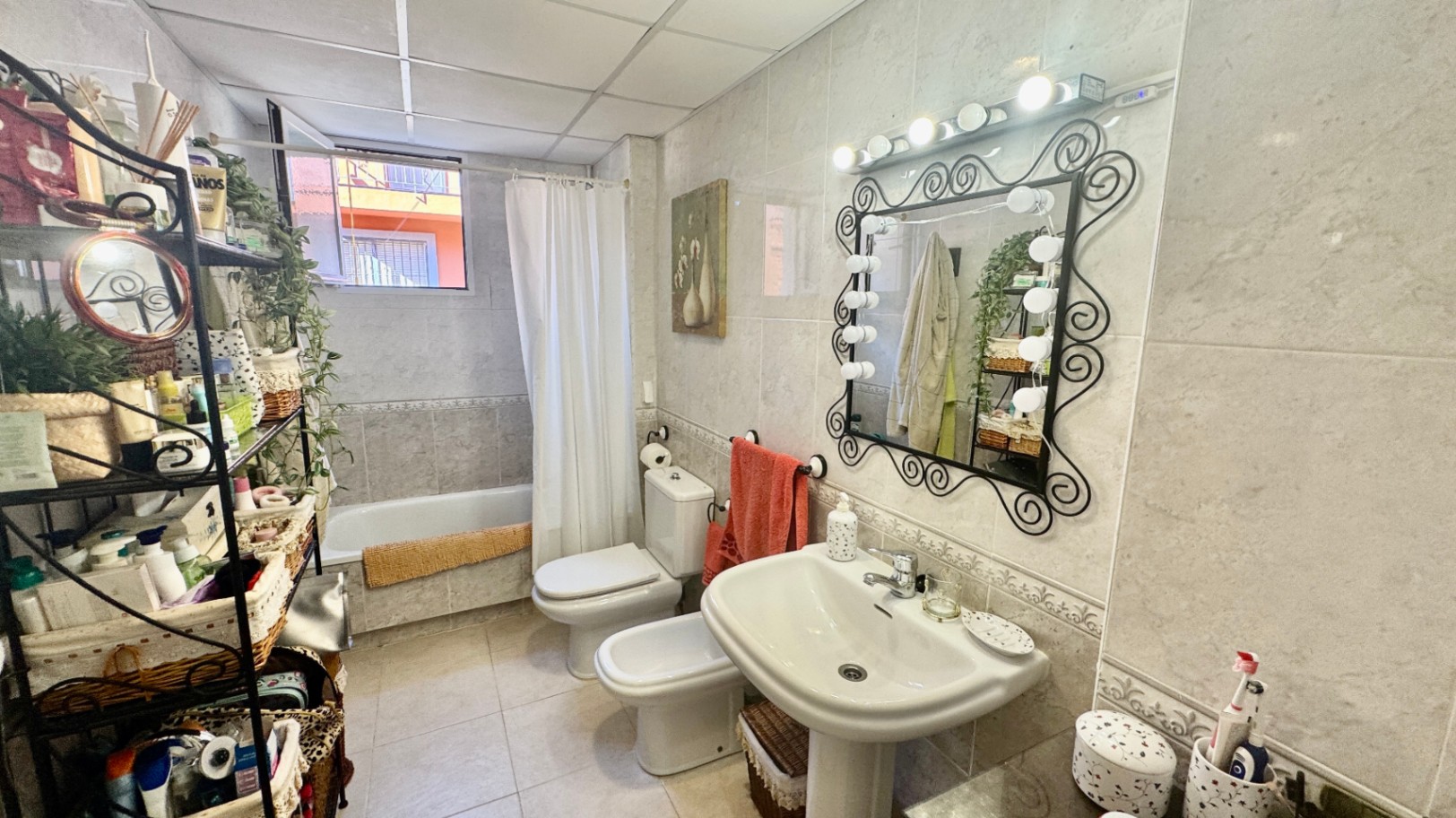 Volledig betegelde badkamer met een kolomwastafel, een spiegel met smeedijzeren frame en theaterverlichting, een bidet en een ligbad.