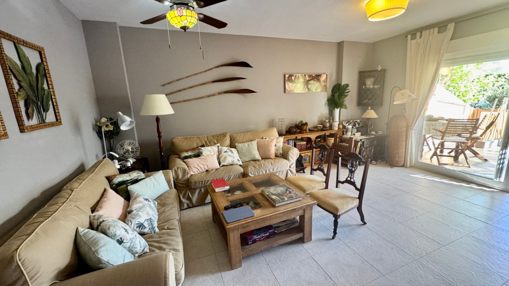 Woonkamer met beige banken, een houten salontafel en glazen deuren die toegang bieden tot een zonnig terras met tuinmeubilair.