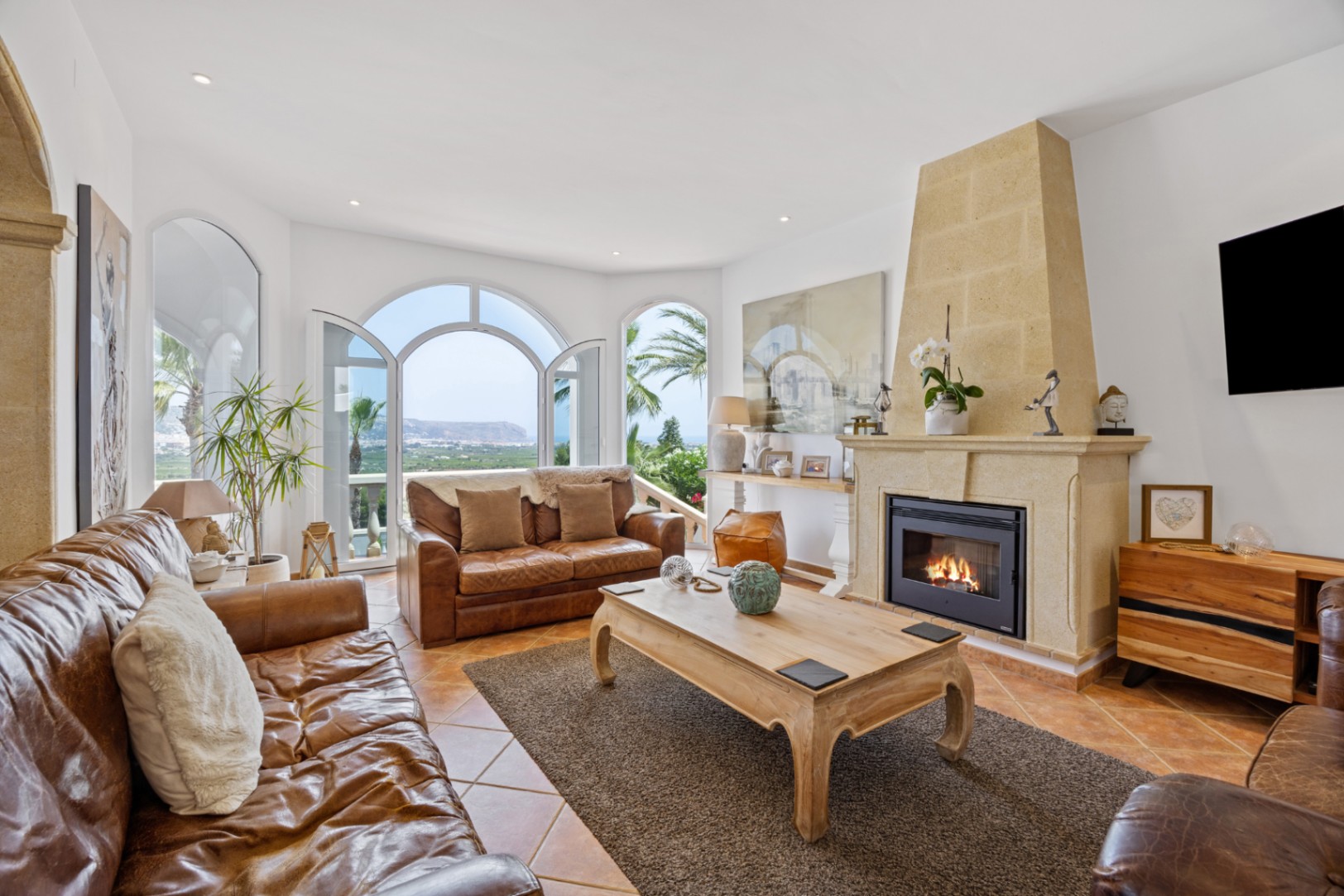 Elegante woonkamer met stenen open haard, lederen banken en boogdeuren die toegang bieden tot een terras met panoramisch uitzicht op zee.