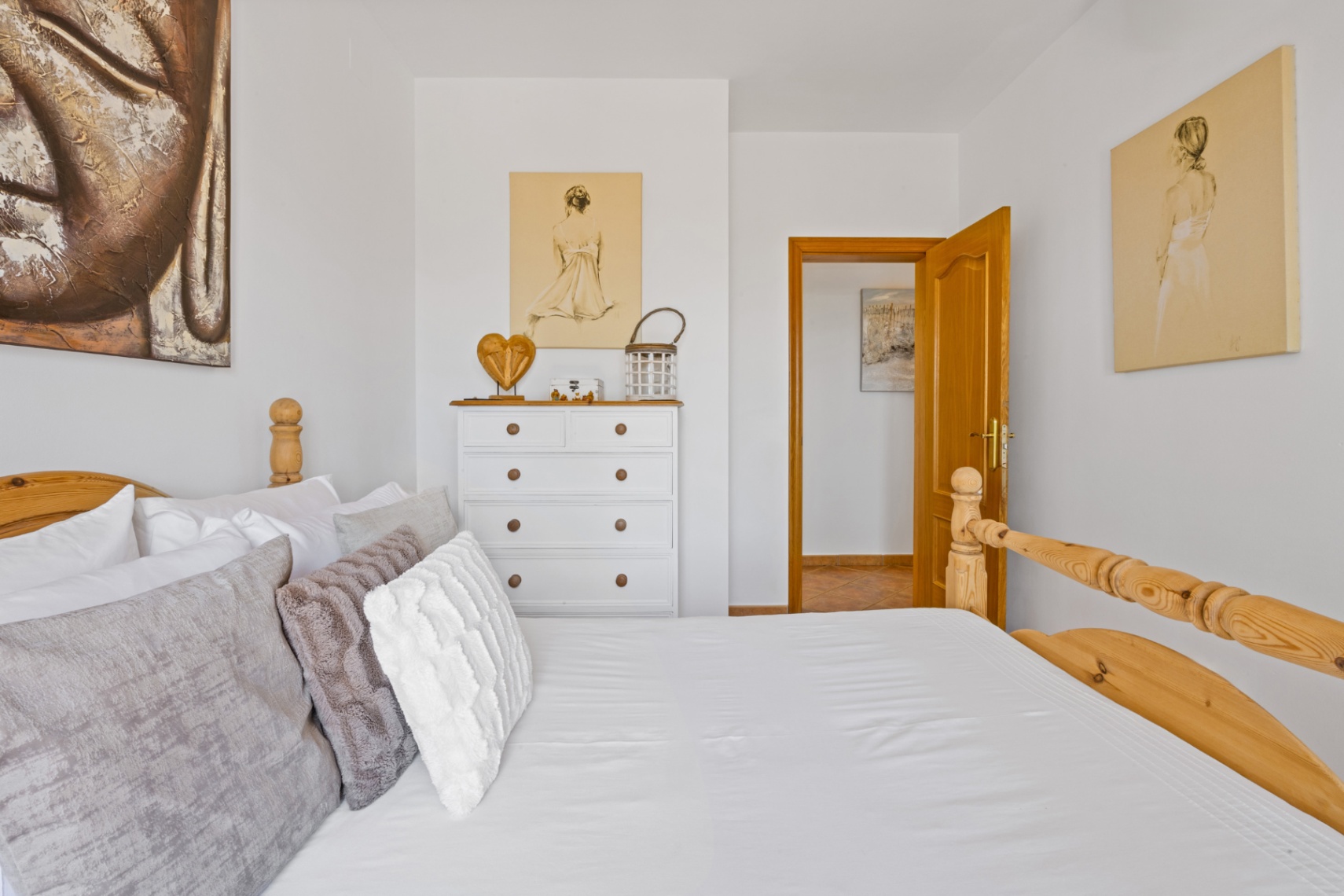 Lichte slaapkamer met massief houten bedframe, witte ladekast met natuurlijke accenten en muren versierd met figuratieve kunst en een groot Boeddha-canvas.