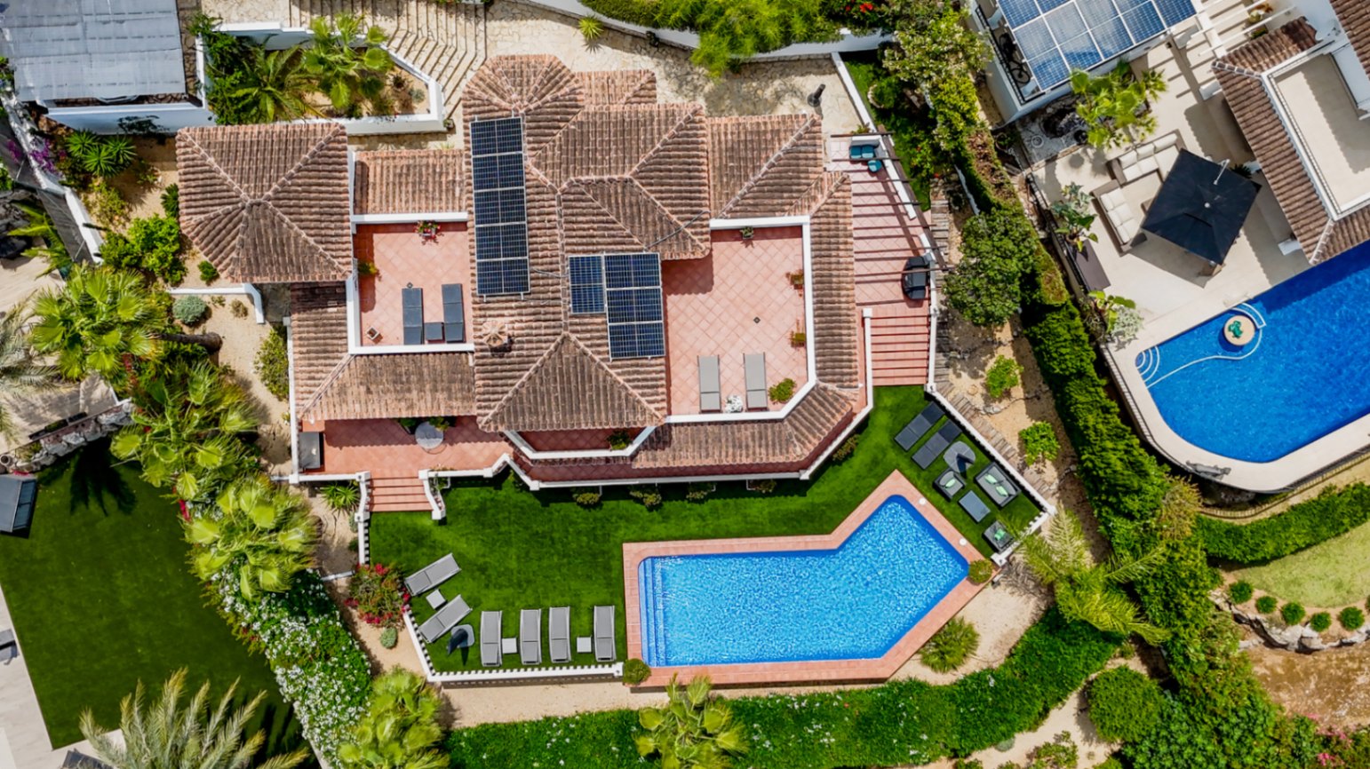 Luchtfoto van een mediterrane villa met een terracotta pannendak, zonnepanelen, diverse zonneterrassen en een hoekig zwembad omgeven door een verzorgd gazon.