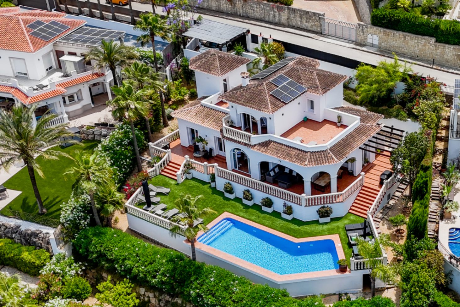 Luchtfoto van een mediterrane villa met geometrisch zwembad, zonnepanelen en terracotta terrassen op meerdere niveaus met witte balustrades.