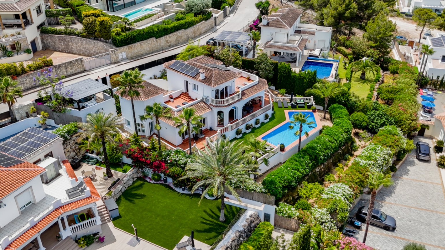 Luchtfoto van een mediterrane villa met terracotta daken, zonnepanelen, een geometrisch privézwembad en aangelegde tuinen met palmbomen.
