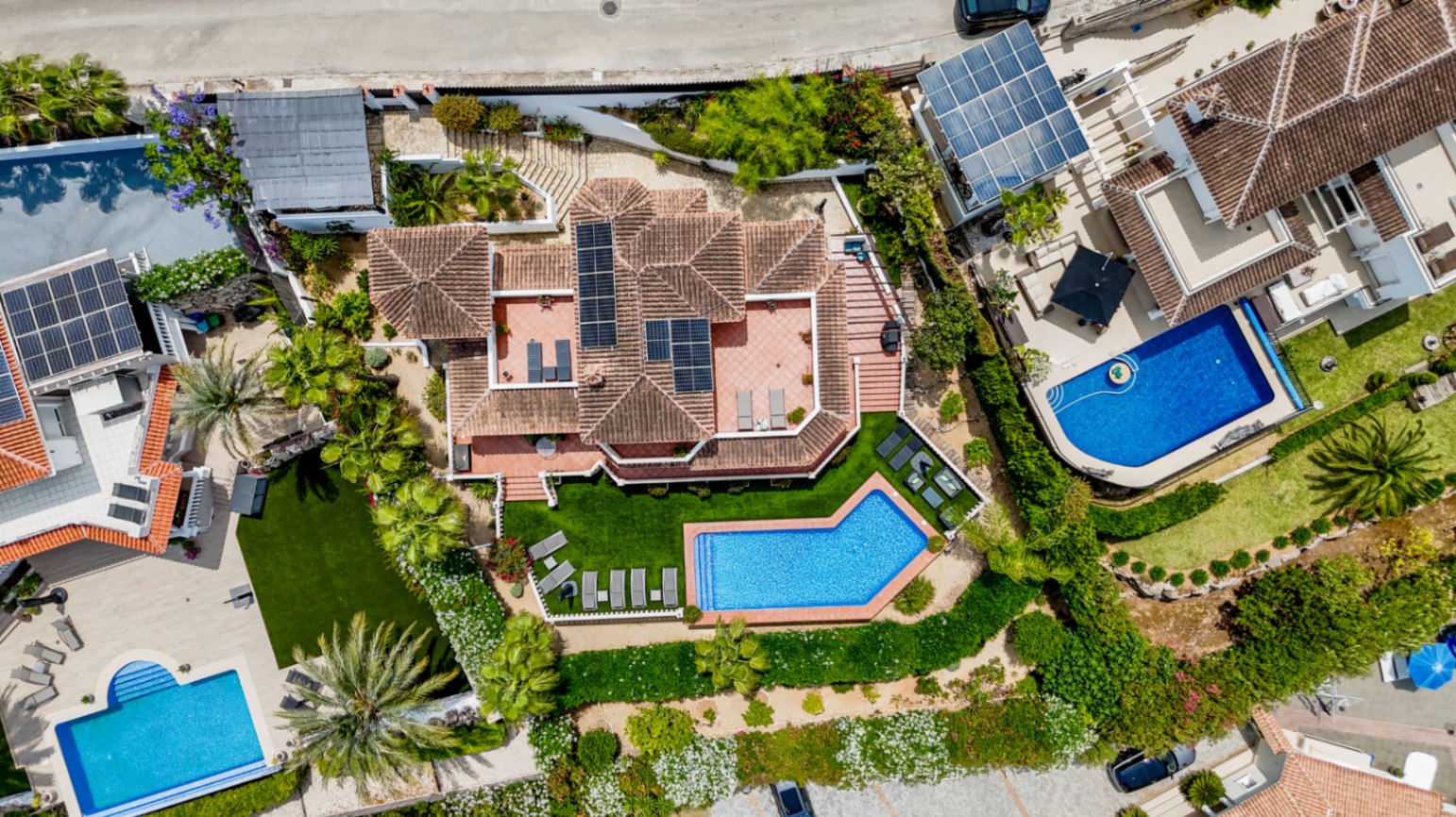 Luchtfoto van een villa met een terracotta dak voorzien van zonnepanelen, een geometrisch zwembad en aangelegde tuinen met palmbomen en zonneterrassen.