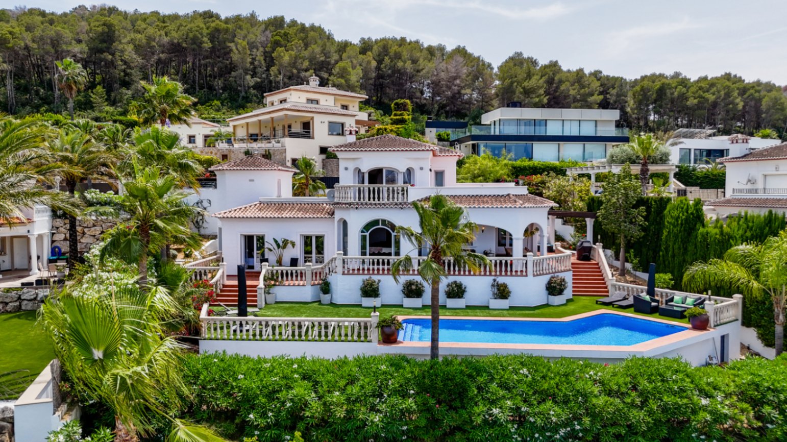 Mediterrane villa met witte gevel, terracotta dak, diverse terrassen en een privézwembad, gelegen in een groene omgeving met palmbomen en pijnbomen.