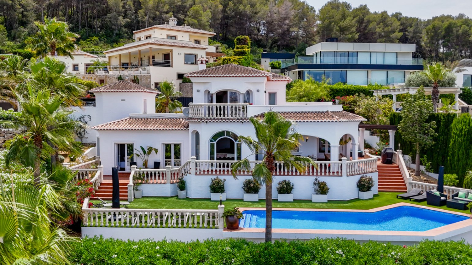 Mediterrane villa met witte gevel, terracotta dakpannen en terrassen met balustrades, uitkijkend op een privézwembad en tuin met palmbomen.