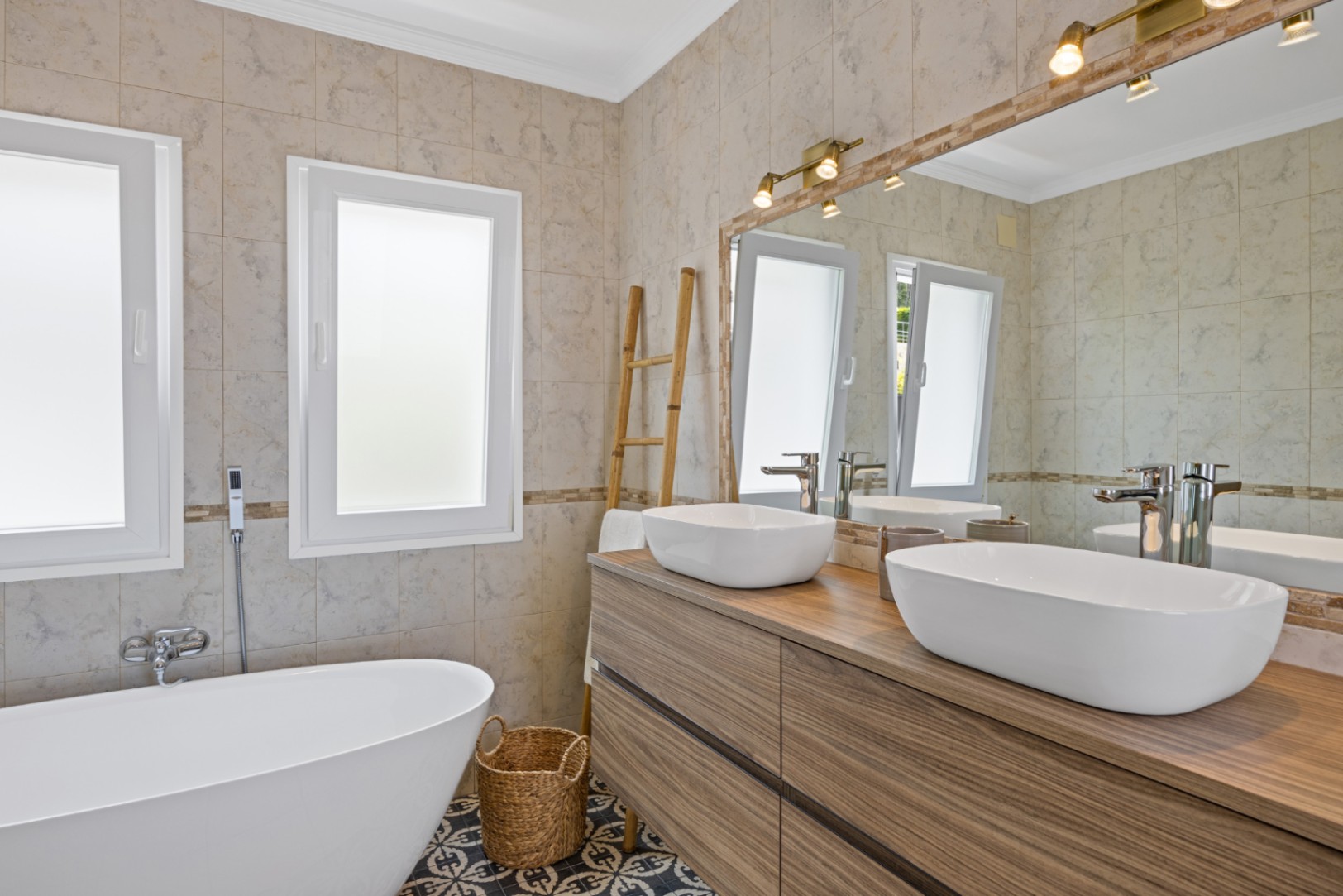 Moderne badkamer met dubbele wastafel op een houten meubel, een vrijstaand bad en gedecoreerde vloertegels, verlicht door spots en natuurlijk licht.