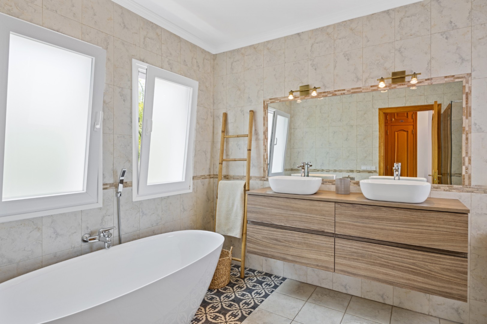 Moderne badkamer met vrijstaand bad, dubbele wastafel op houten meubel en patroontegels. Grote spiegel met messing armaturen en ramen met melkglas.