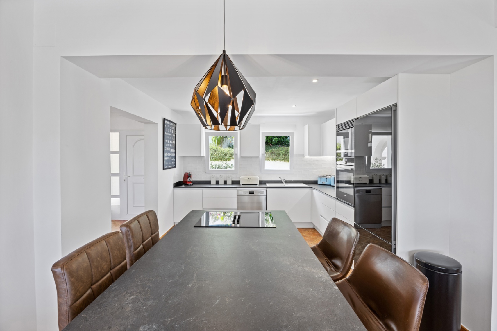 Moderne keuken met een groot donkerstenen kookeiland, geïntegreerde inductiekookplaat, bruine lederen stoelen en witte kasten met inbouwapparatuur.