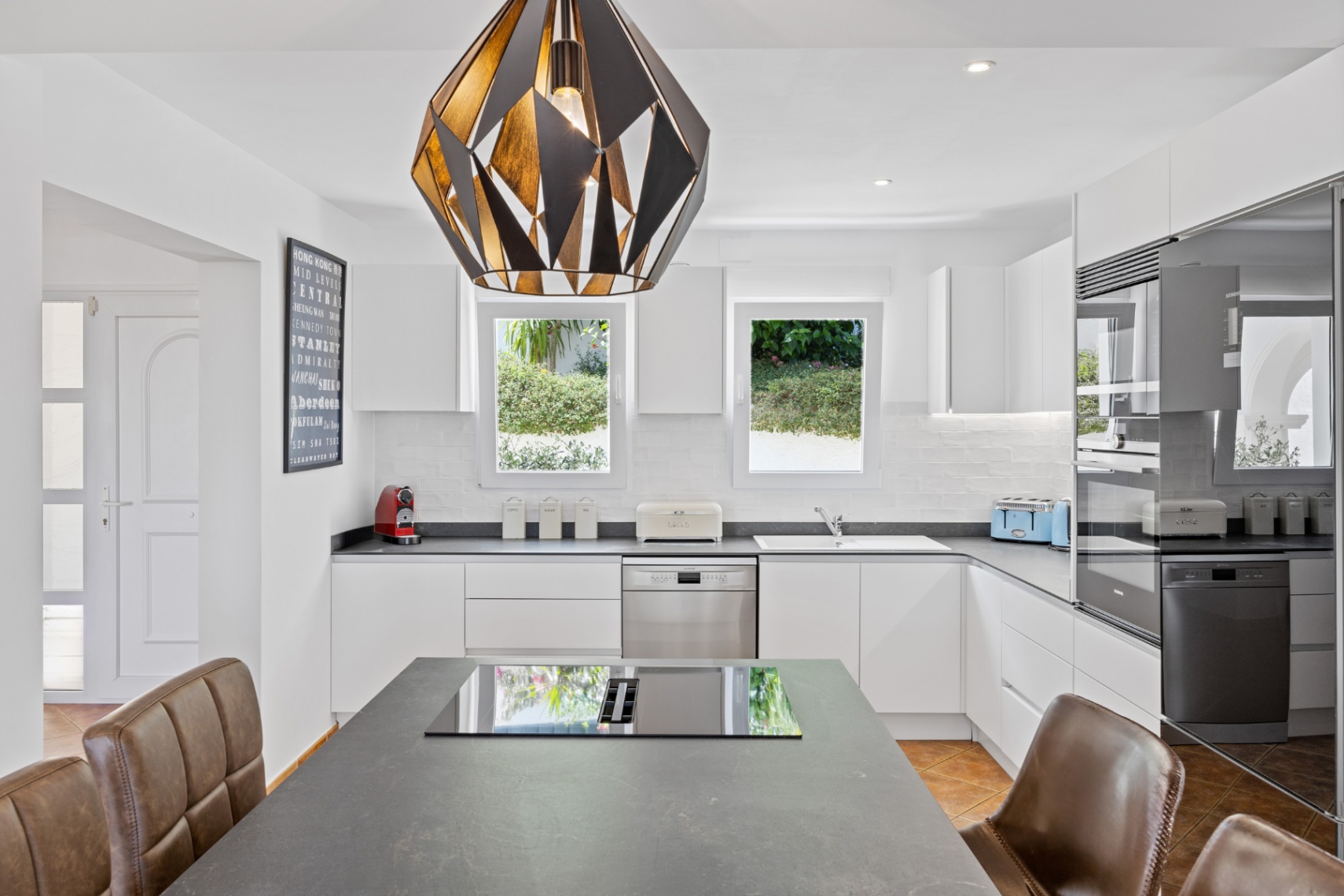 Moderne keuken met greeploze witte kasten, donkere stenen werkbladen en een kookeiland met inductiekookplaat onder een opvallende geometrische hanglamp.