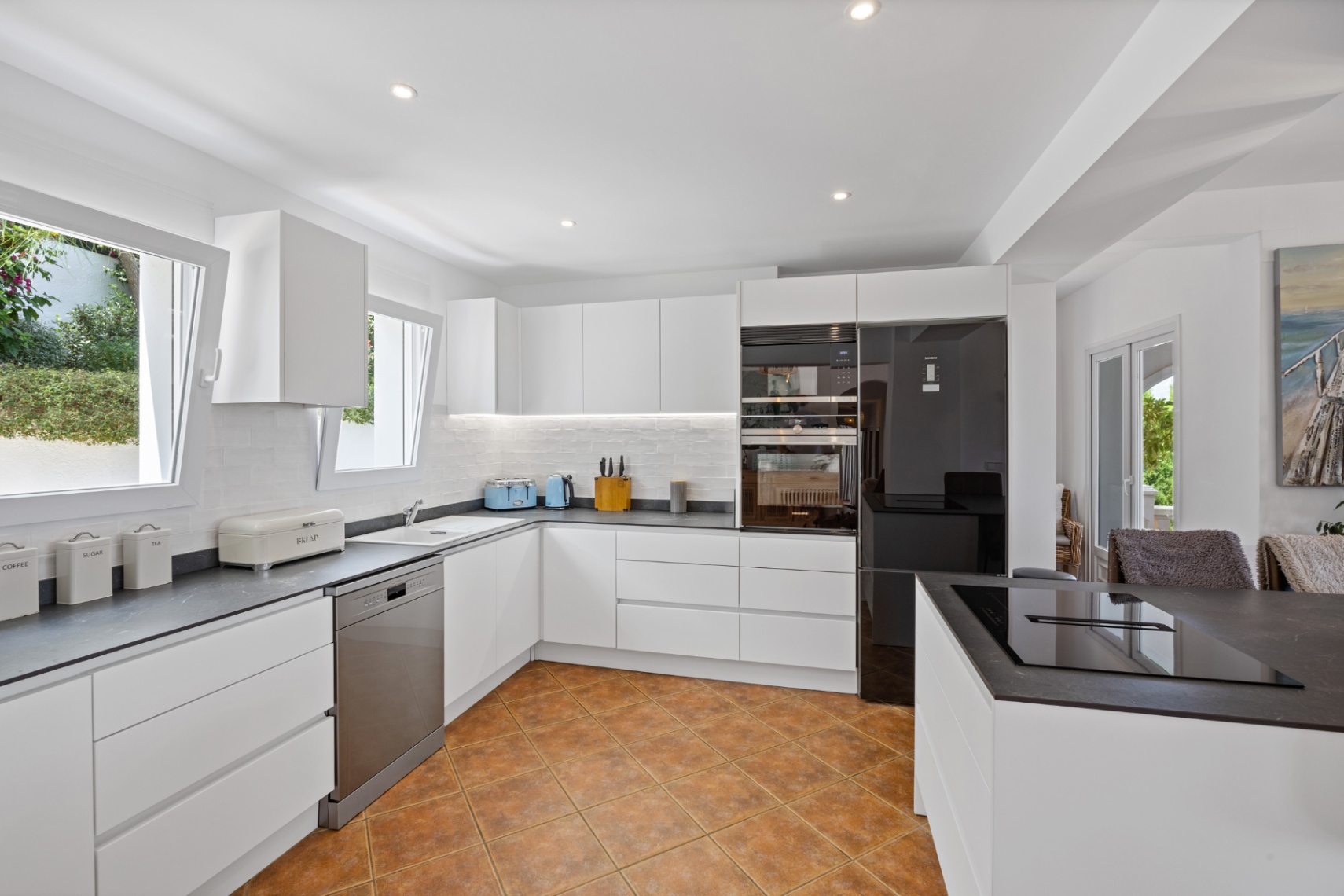 Moderne keuken met greeploze witte kasten, donkere werkbladen met steeneffect, inbouwapparatuur en een terracotta-stijl keramische vloer.