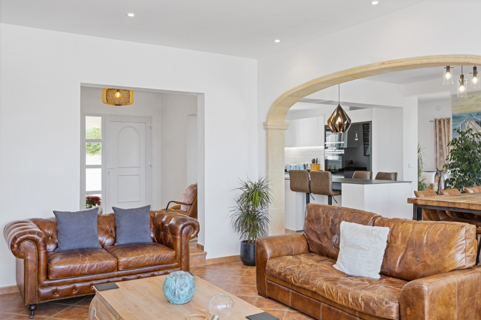 Open woonkamer met lederen banken, terracotta tegels en een stenen boog die toegang biedt tot een moderne keuken met ontbijtbar.