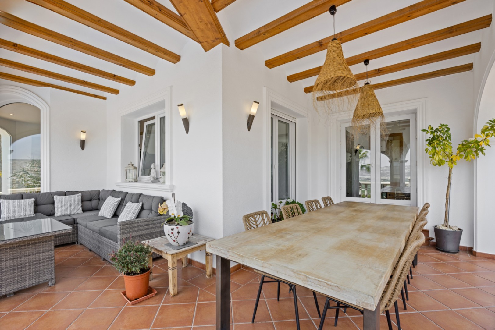 Overdekt terras met houten balken, terracotta vloer, een robuuste houten eettafel met gevlochten stoelen en een grijze wicker loungehoek.