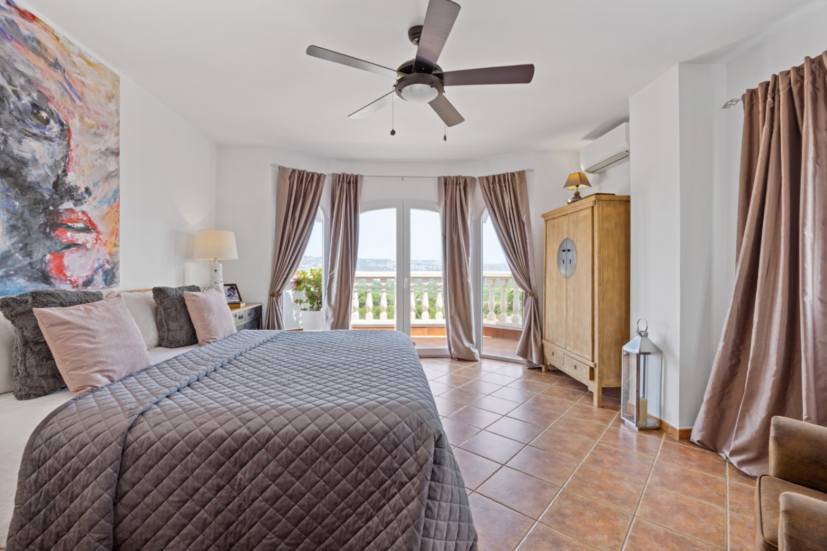 Ruime slaapkamer met terracotta vloertegels, een plafondventilator en boogdeuren die toegang bieden tot een balkon met uitzicht op het landschap.