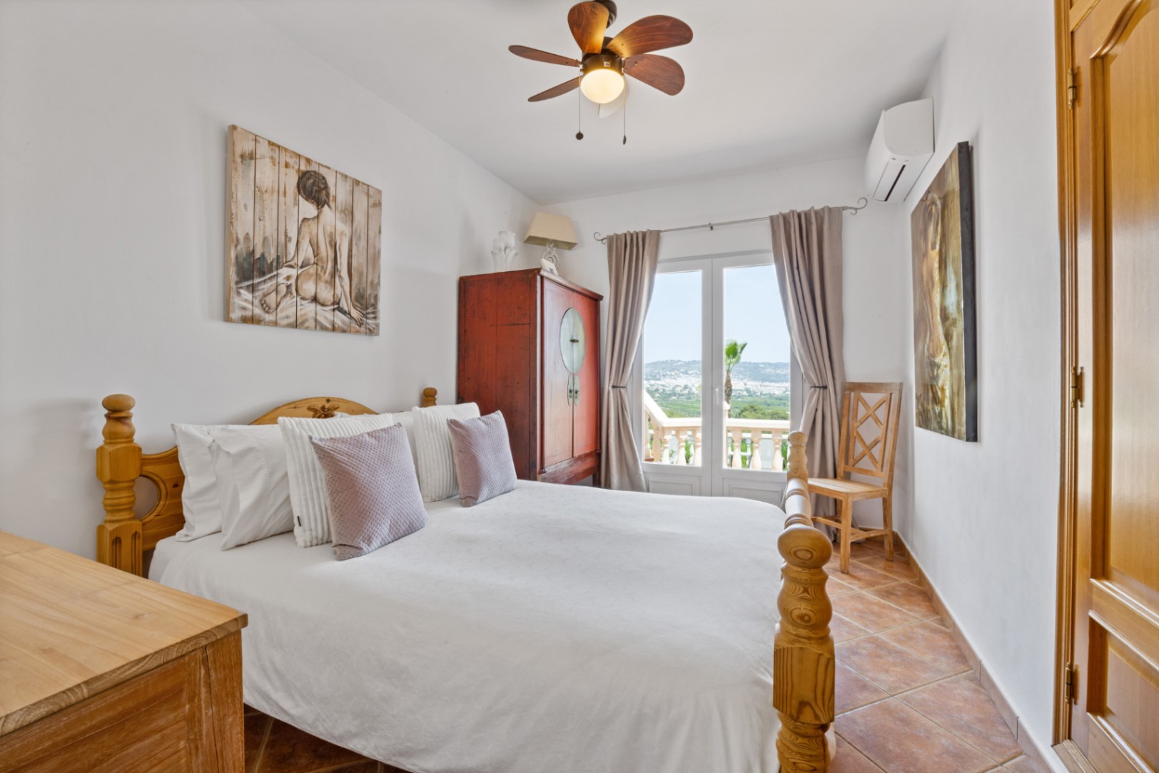 Slaapkamer met houten bed, terracotta tegelvloer en openslaande deuren naar een balkon met uitzicht. Inclusief plafondventilator en airconditioning.