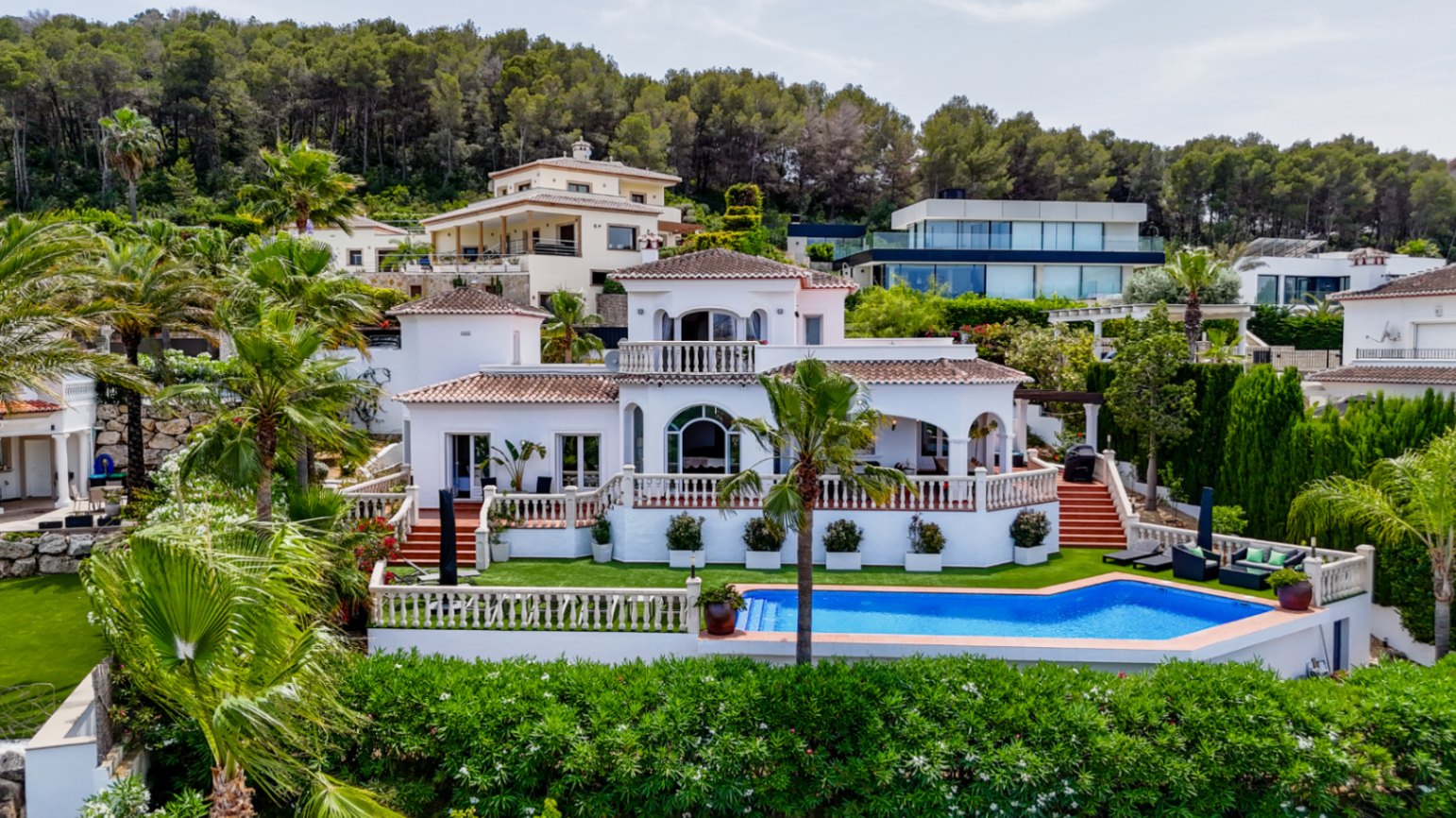 Verhoogd zicht op een witte mediterrane villa met terracotta daken, boogvensters en een privézwembad omgeven door palmbomen en dennenbos.