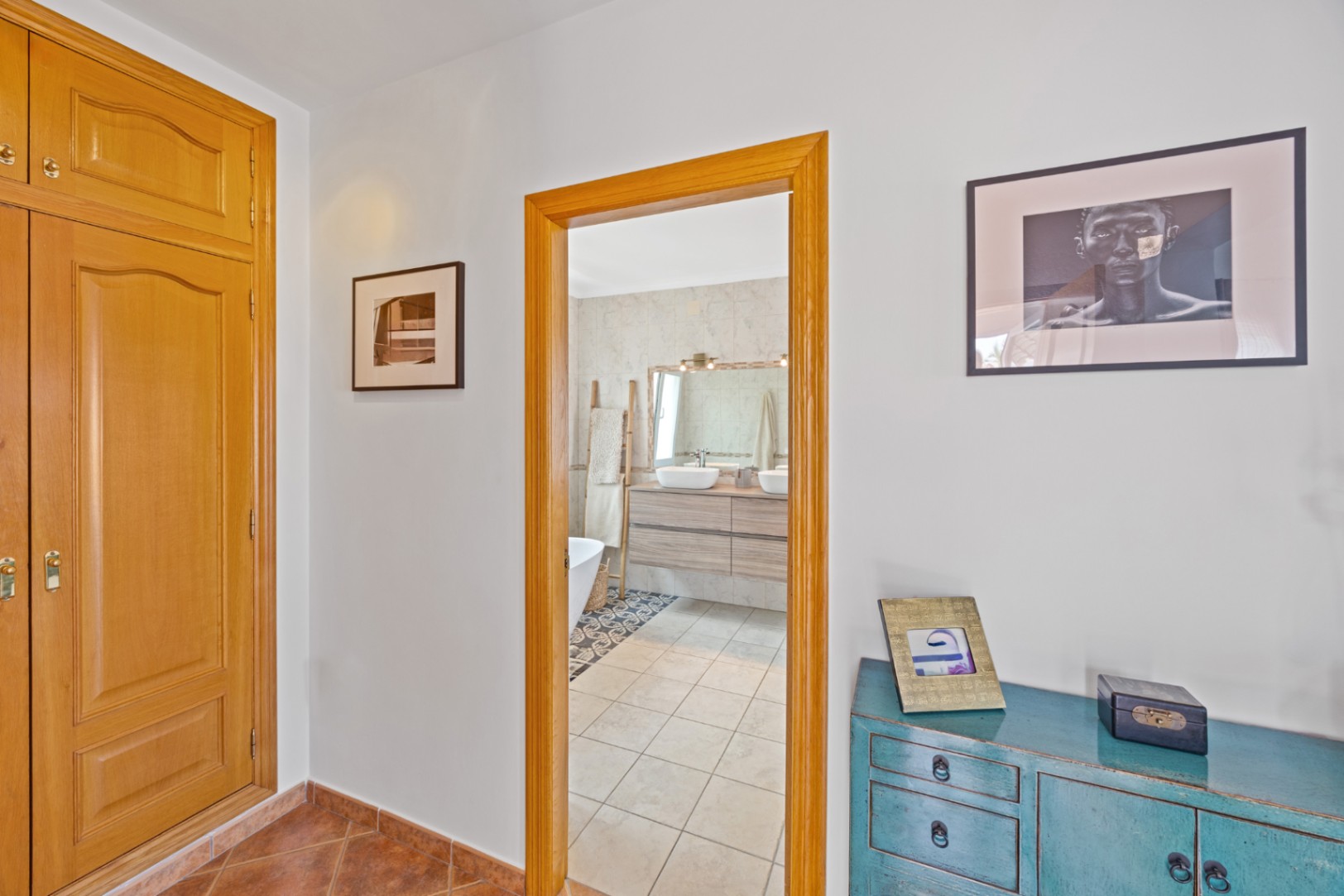 Zicht vanuit de gang met houten inbouwkasten en een blauw dressoir op een badkamer met dubbele wastafel, houten meubel en betegelde vloer.