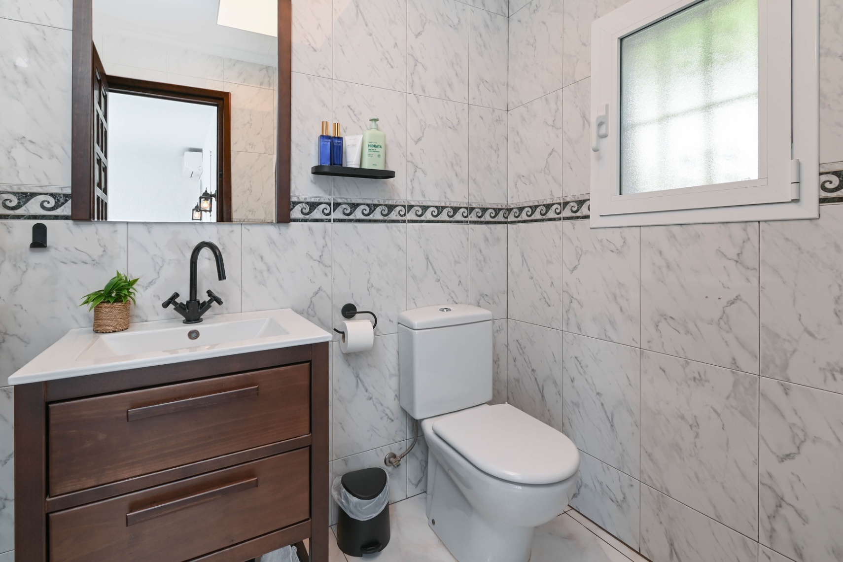 Badkamer met marmerlook wandtegels, een donkerhouten wastafelmeubel, matzwarte kranen en een klein raam met melkglas voor natuurlijke lichtinval.