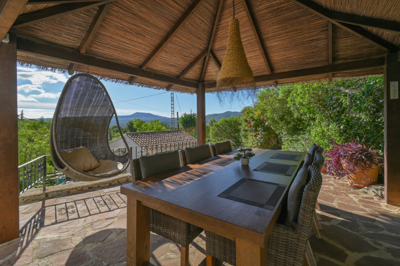 Buiten-eethoek onder een rieten gazebo met een houten tafel, rotan stoelen en een hangstoel met uitzicht op de omliggende bergen.