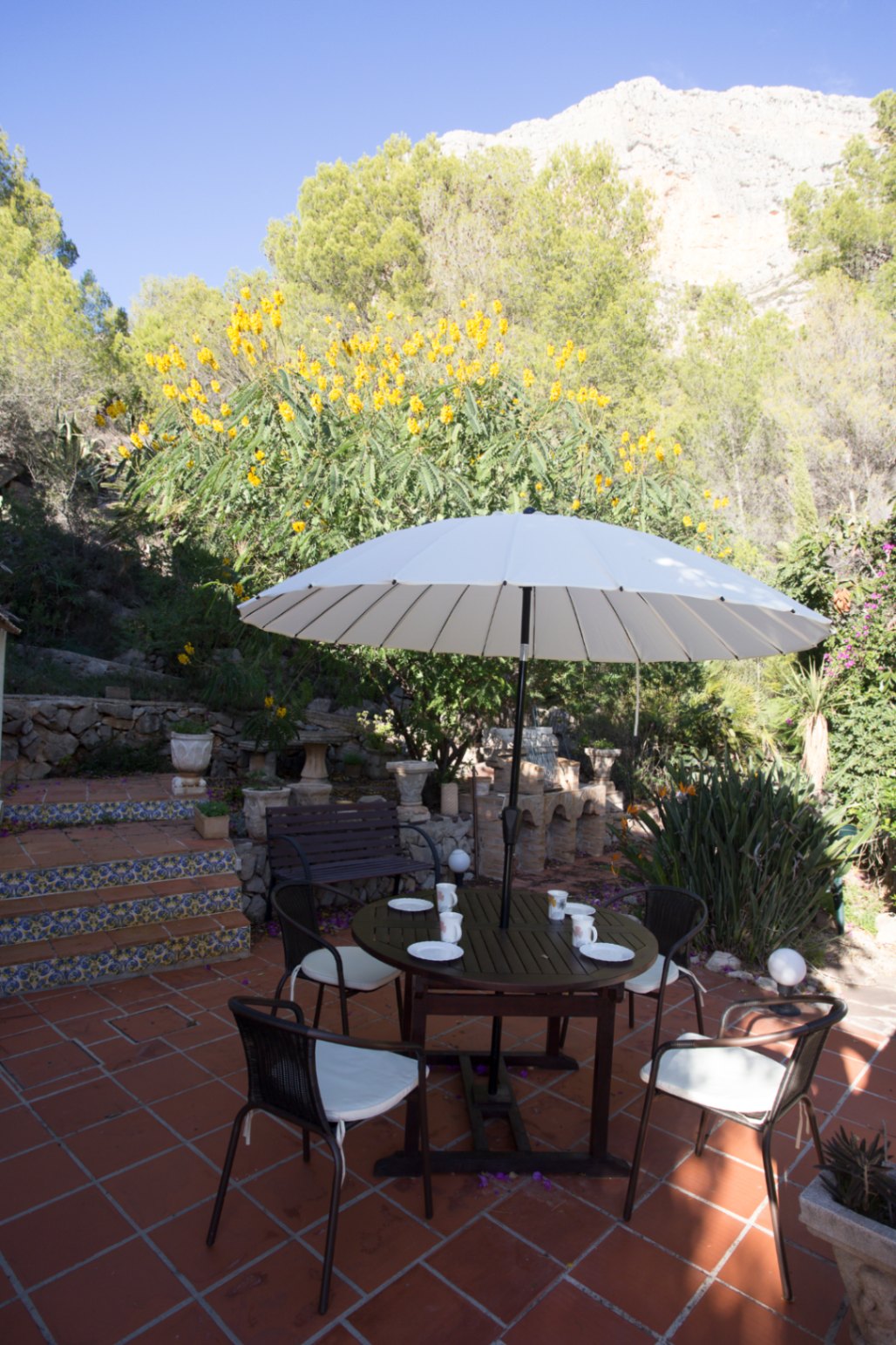 Buitenterras met terracotta tegels, een ronde eettafel onder een witte parasol en decoratieve betegelde trappen, gelegen tegen een achtergrond van bergen en groen.