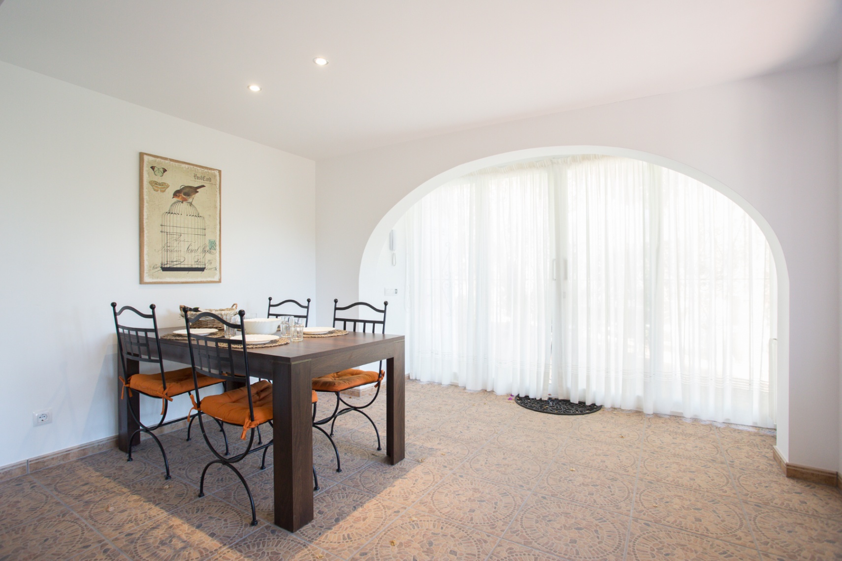 Eethoek met donkerhouten tafel en smeedijzeren stoelen. Een grote boogvormige doorgang met witte gordijnen verlicht de karakteristieke patroontegelvloer.