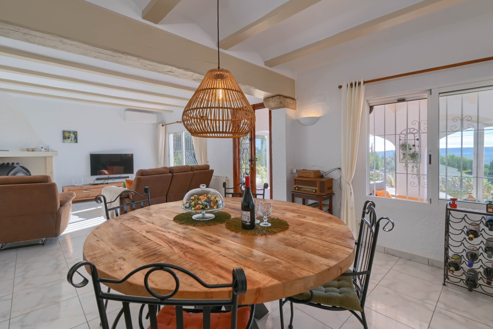 Eethoek met een ronde houten tafel, smeedijzeren stoelen en een rieten hanglamp, onder een plafond met zichtbare balken grenzend aan de woonkamer.