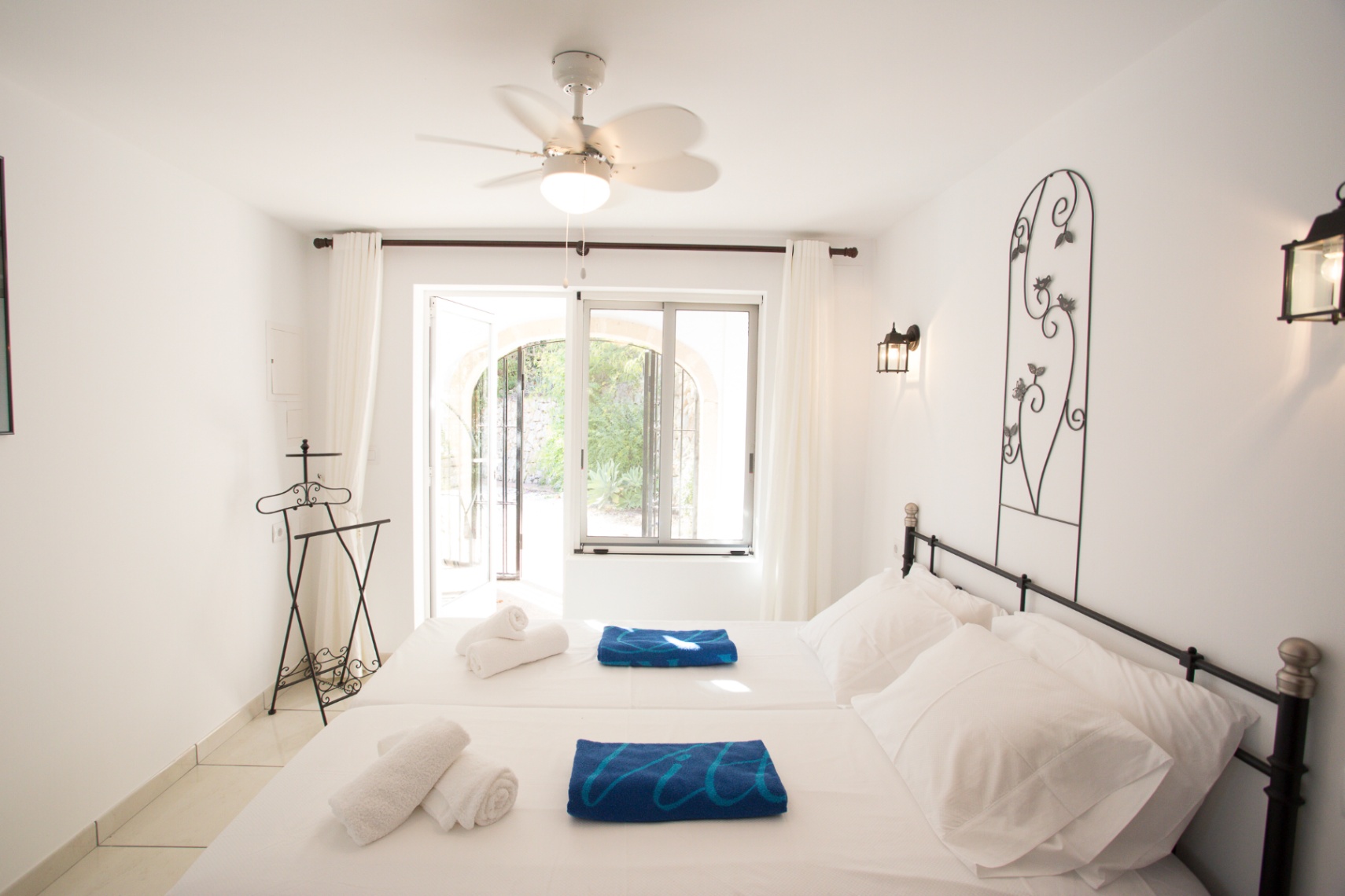 Lichte slaapkamer met witte muren, een smeedijzeren hoofdbord en plafondventilator. Openslaande deuren geven toegang tot een zonnig terras en de tuin.