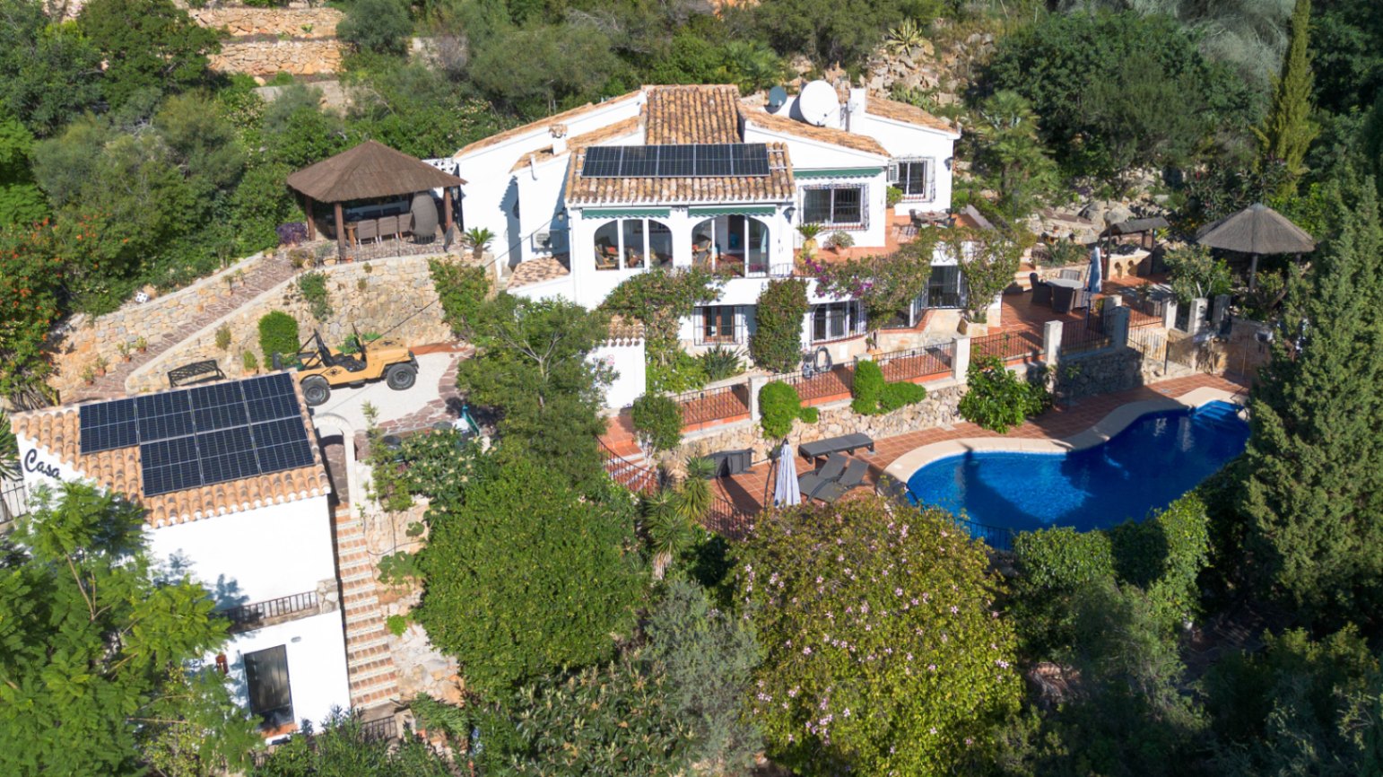 Luchtfoto van een mediterrane villa met zonnepanelen op terracotta daken, een niervormig zwembad en terrasvormige tuinen met natuurstenen muren.