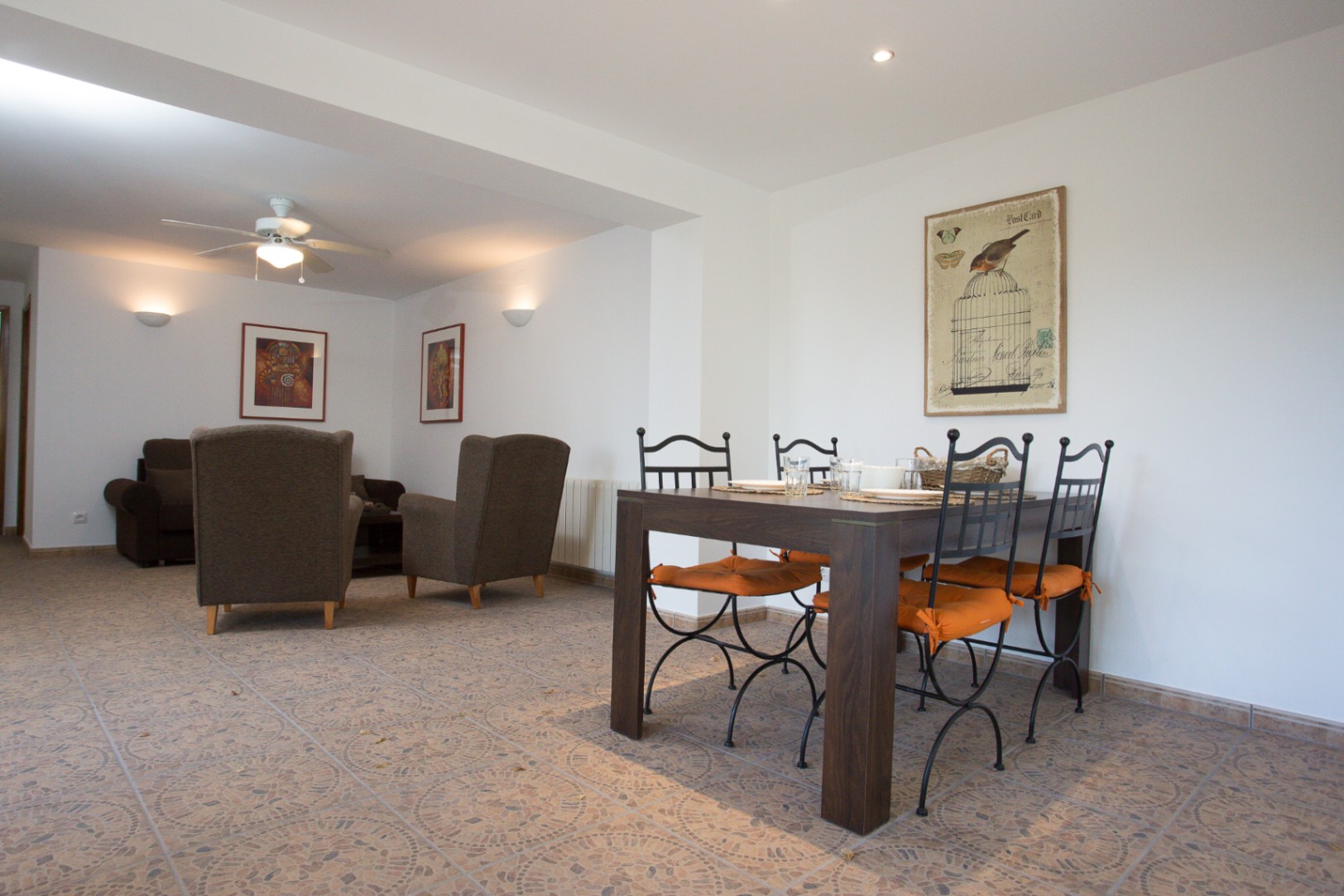 Open woon- en eetkamer met een donkerhouten eettafel, smeedijzeren stoelen en oranje kussens. De ruimte is voorzien van een gedessineerde tegelvloer en een zithoek.