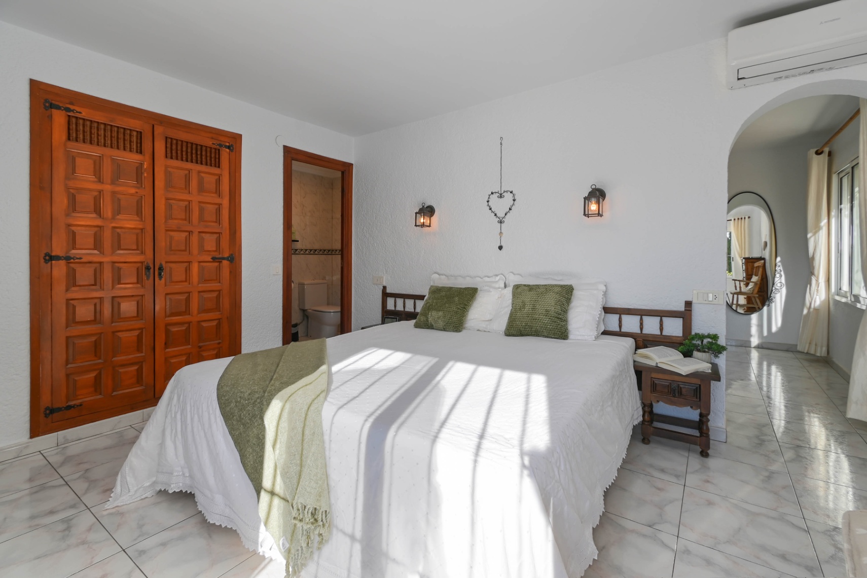 Slaapkamer met een gesneden houten inbouwkast, lichte marmeren vloer en badkamer en suite. Natuurlijk licht valt op het bed met groene accenten en wandlampen in lantaarnstijl.
